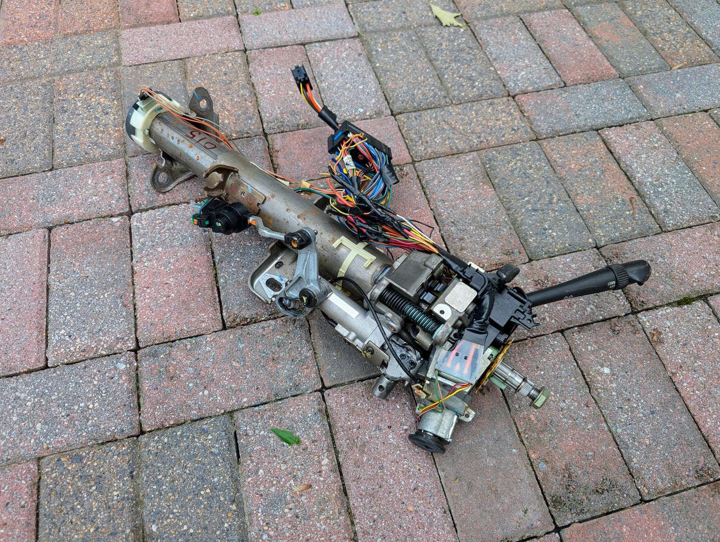 A used General Motors OEM Steering Column for 2000-2002 GM Full Size Trucks with visible wiring, cables, and connectors is shown resting on a brick pavement.