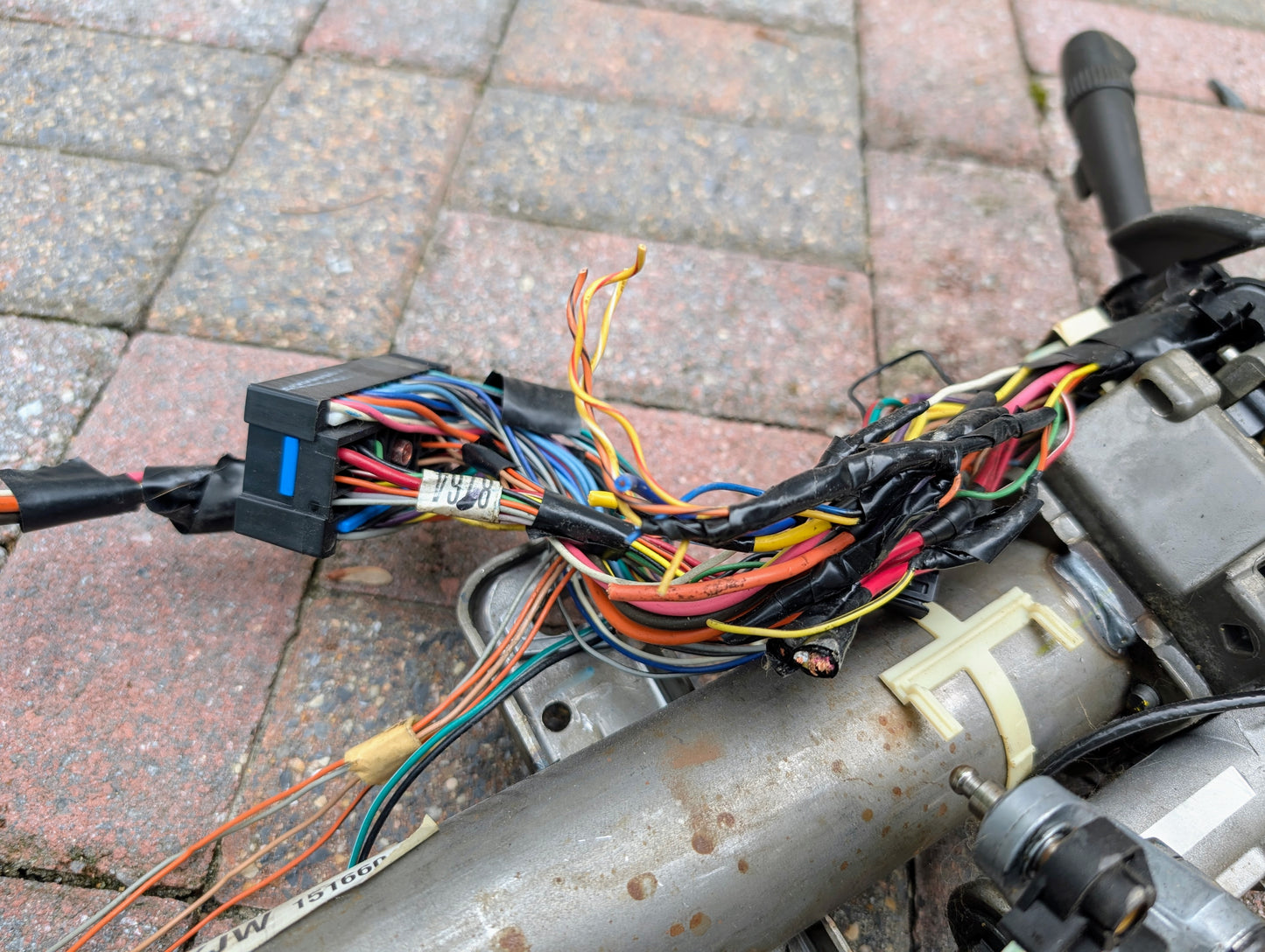 A close-up of multicolored wires, some wrapped in black tape, connected to a General Motors OEM Steering Column for 2000-2002 GM Full Size Trucks, resting on a brick-patterned surface.