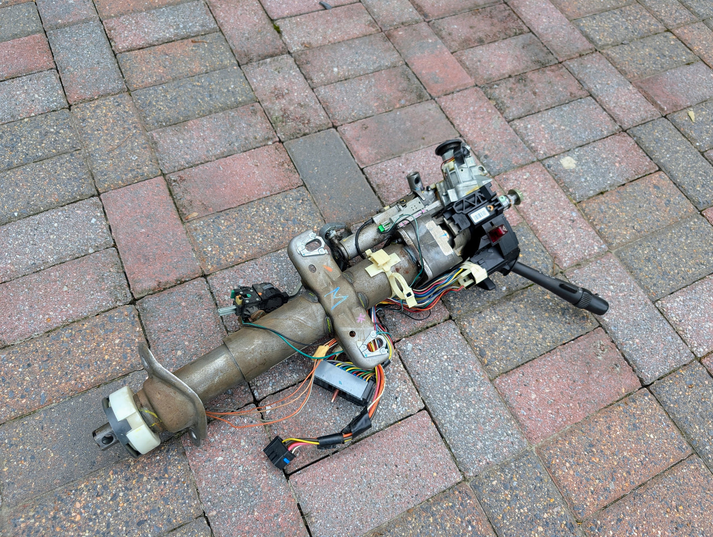 A disassembled General Motors OEM Steering Column for 2000-2002 GM Full Size Trucks, with wiring and electronic components for automatic transmission, rests on red and gray brick pavement.