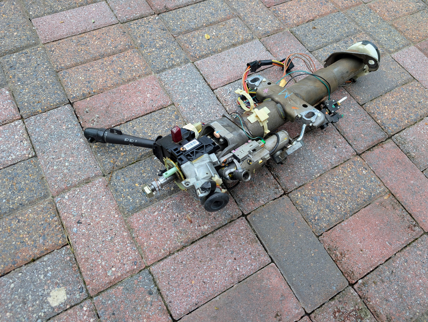 An OEM Steering Column for 2000-2002 GM Full Size Trucks by General Motors, complete with wiring and control levers, is pictured disassembled on a brick-paved surface.