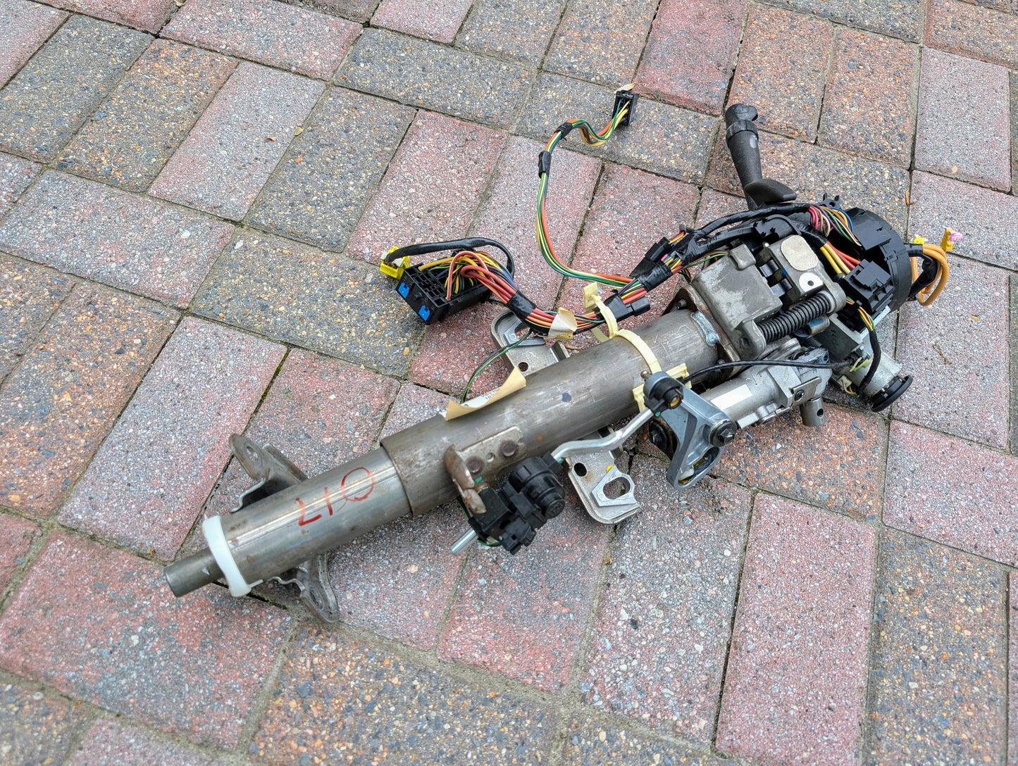 A disassembled General Motors OEM Steering Column for 2003-2006 GM Full Size Trucks, with wiring harnesses, from a model with automatic transmission, is shown on a red and gray brick surface.