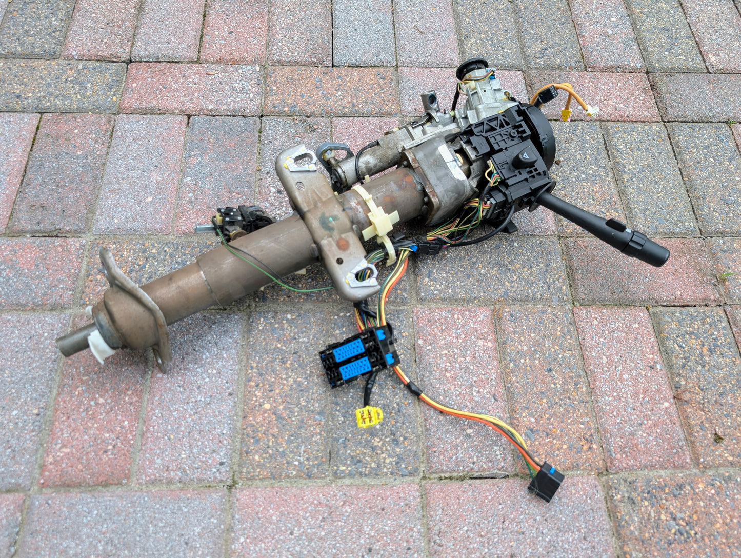 A General Motors OEM Steering Column for 2003-2006 GM Full Size Trucks with wiring and controls attached is placed on a paved surface made up of rectangular gray and red bricks.