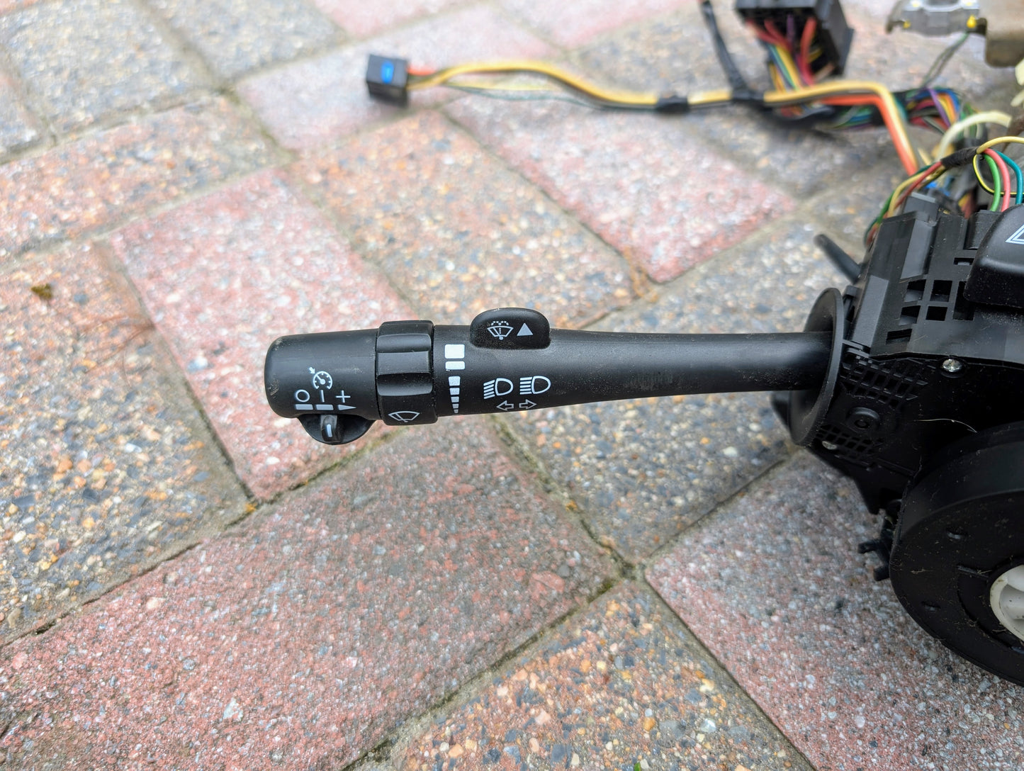 A close-up of a General Motors OEM Steering Column for 2003-2006 GM Full Size Trucks shows the windshield wiper and headlight control stalk with symbols, placed on a paved surface among connected wires and components.