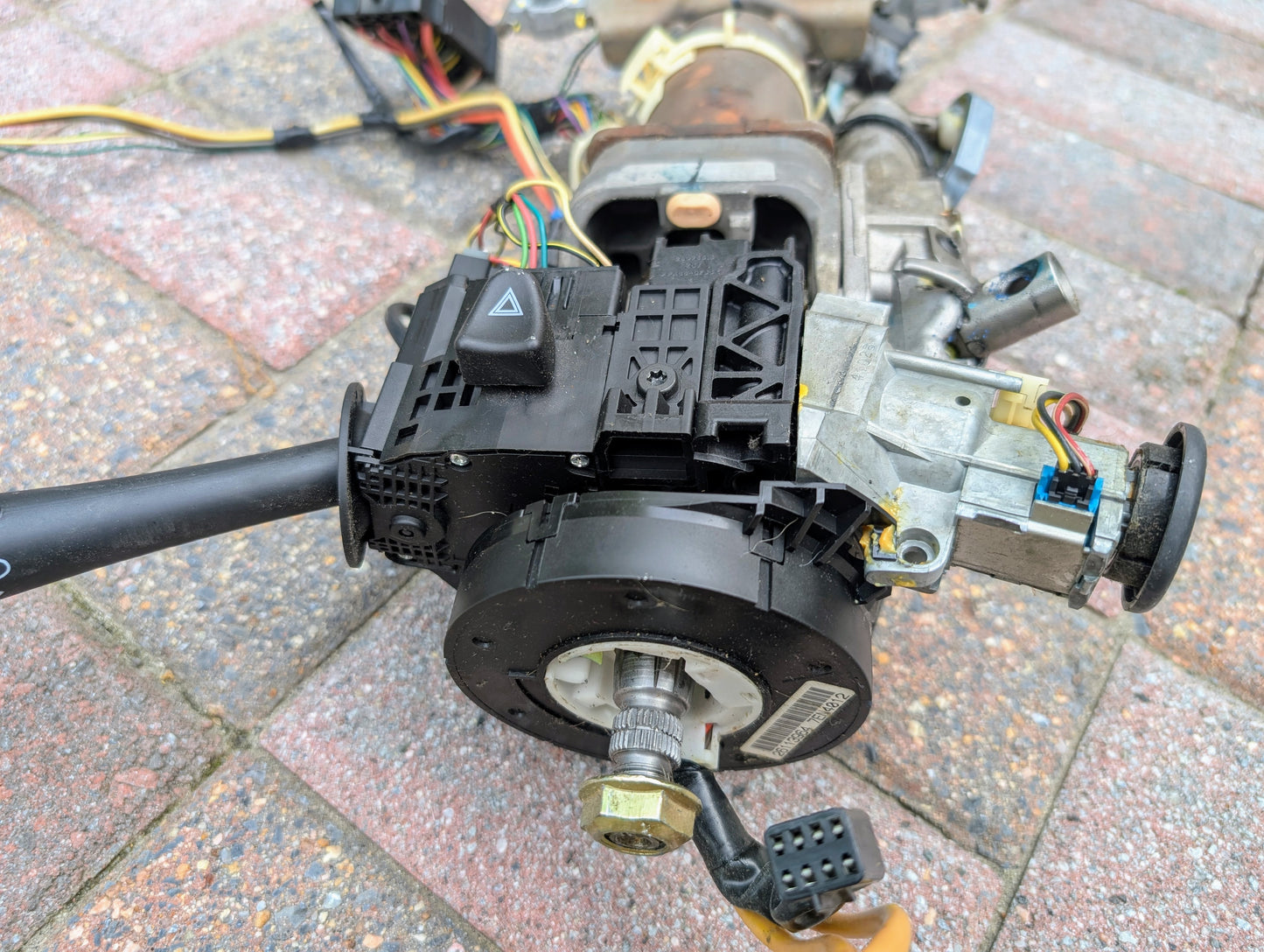 A close-up of a General Motors OEM Steering Column for 2003-2006 GM Full Size Trucks with automatic transmission, displaying wiring, connectors, and mechanical parts on brick. The hazard light switch and turn signal stalk are visible.
