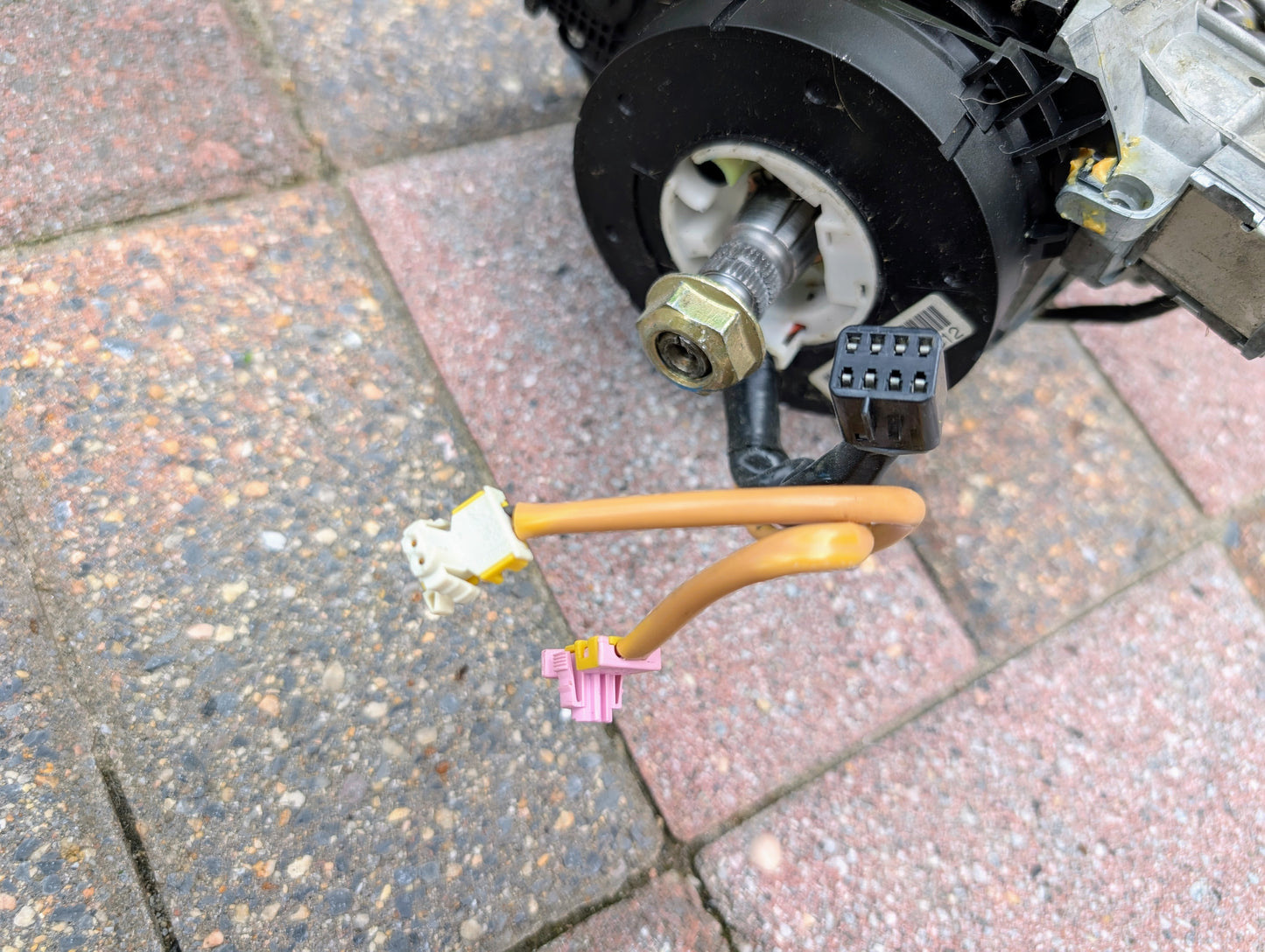 Close-up of electrical connectors and cables attached to a General Motors OEM Steering Column for 2003-2006 GM Full Size Trucks, shown resting on multicolored paving stones.