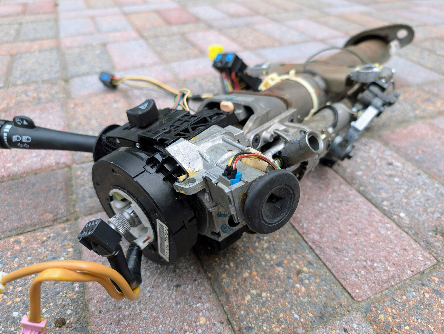 A close-up of a disassembled General Motors OEM Steering Column for 2003-2006 GM Full Size Trucks, showing exposed wiring and parts, laid out on a paved brick surface outdoors.