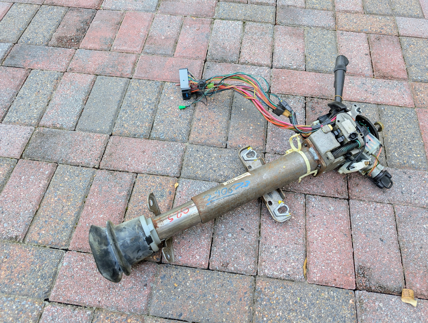 A used General Motors OEM Steering Column w/Tilt, Cruise Control, and Console Shift for 1998-2005 Chevy S10 Blazer lies on brick pavement. It has attached wires, a lever, colored writing, and visible signs of use.