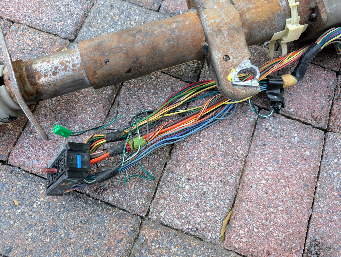 A General Motors OEM Steering Column with tilt, cruise control, and console shift for 1998-2005 Chevy S10 Blazer rests next to a rusty pipe on red and brown brick pavers.