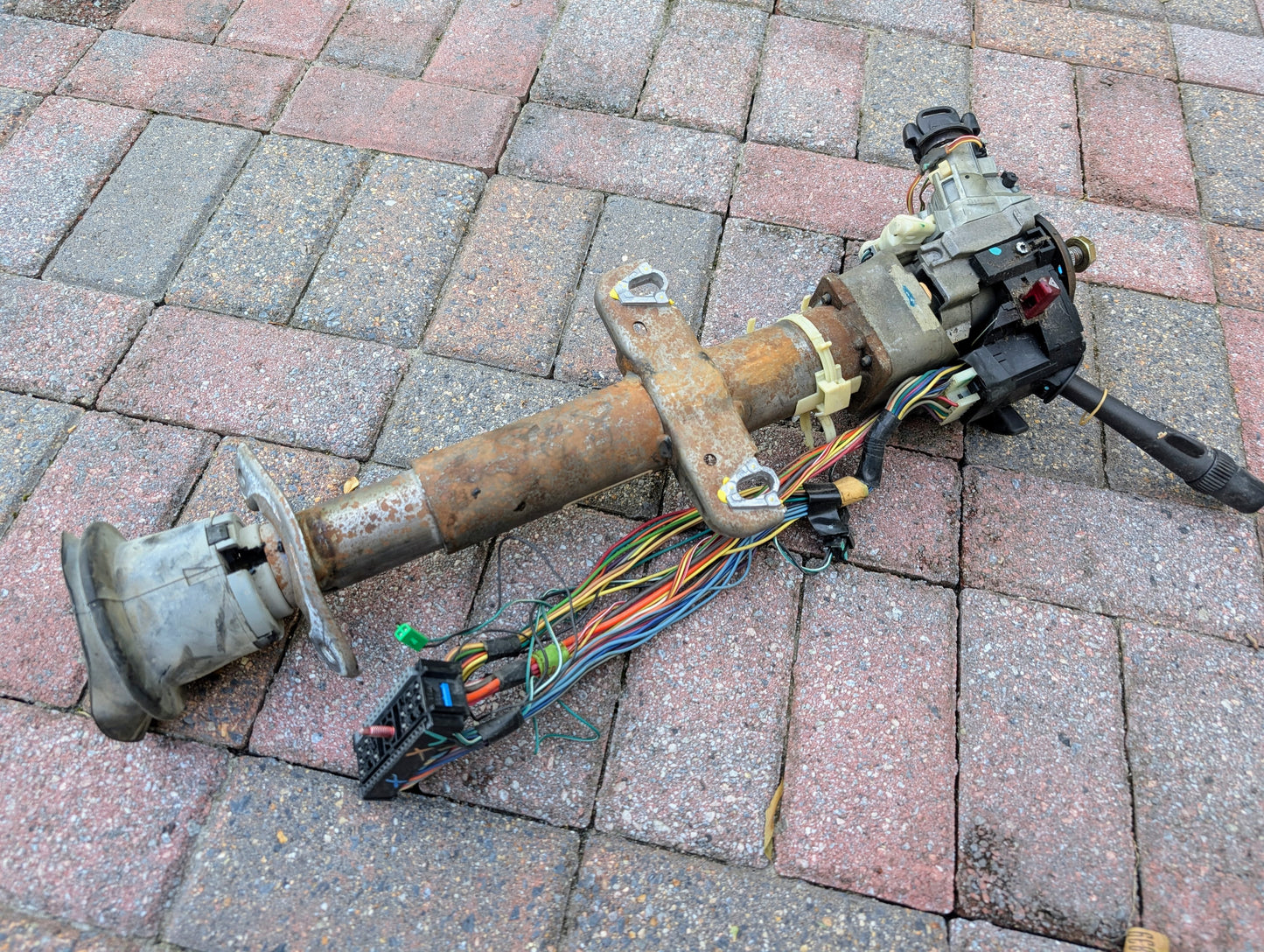 A General Motors OEM Steering Column with tilt, cruise control, and console shift for 1998-2005 Chevy S10 Blazer and more, complete with wiring and connectors, is shown resting on red and gray brick pavement.