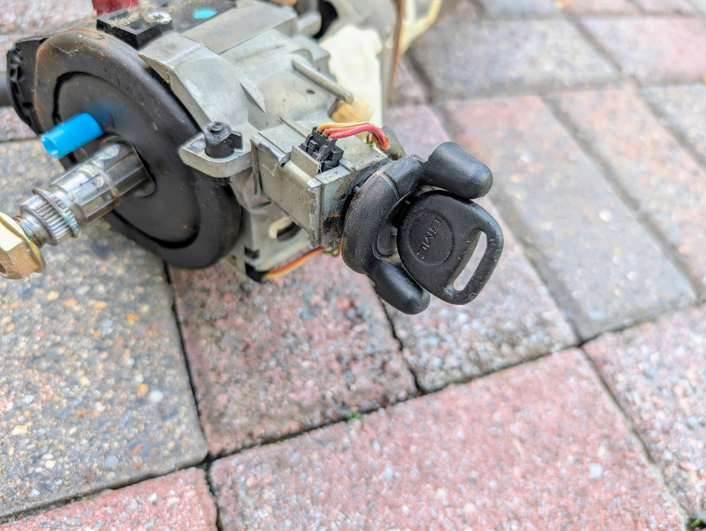 A General Motors OEM steering column with tilt, cruise control, and console shift for 1998-2005 Chevy S10 and Blazer is shown close up on red and gray paving stones, featuring the ignition switch with key inserted.