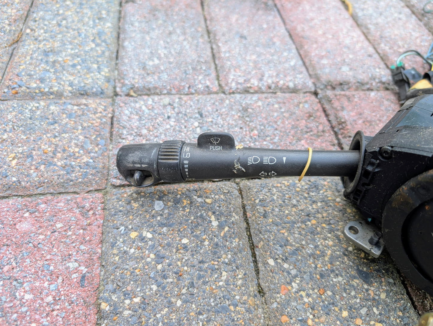 A used General Motors OEM Steering Column with tilt, cruise control, and console shift for 1998-2005 Chevy S10 Blazer and more lies on textured brick, showing slight wear. Black with white symbols and a PUSH button.