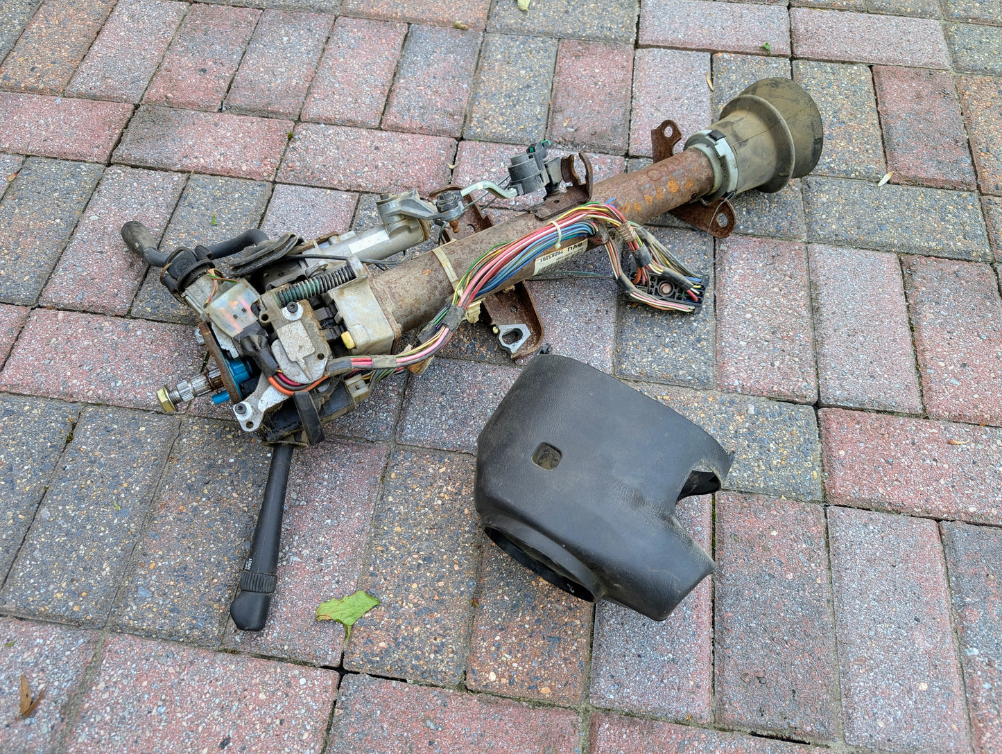 A used General Motors OEM Steering Column with Tilt, Cruise Control, and Column Shift for 1998-2005 Chevy S10 Blazer lies on pavement with attached wiring and a detached black plastic cover.
