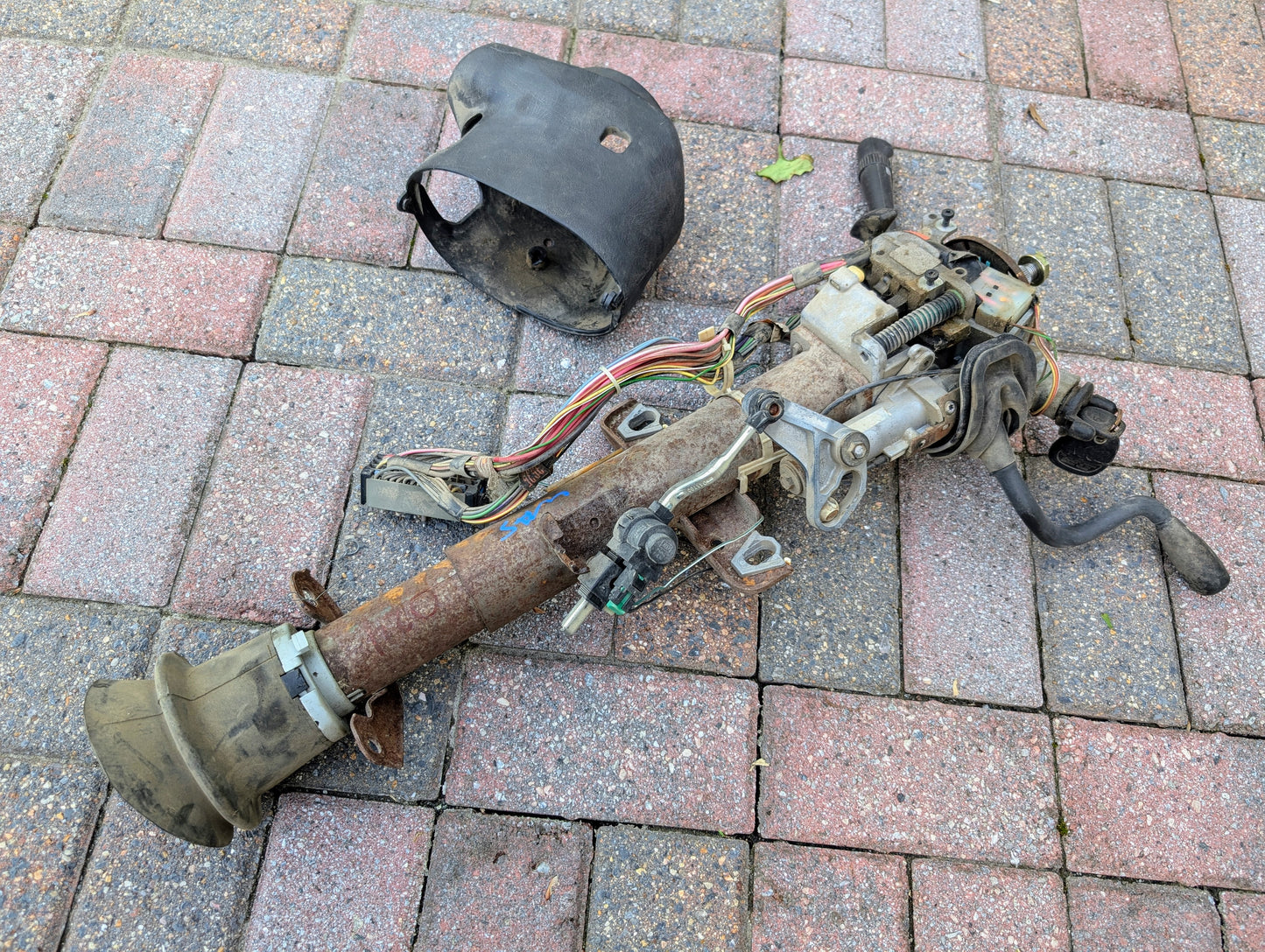 A General Motors OEM Steering Column with Tilt, Cruise Control, and Column Shift for 1998-2005 Chevy S10/Blazer lies disassembled with wiring and covers on a tiled outdoor surface.