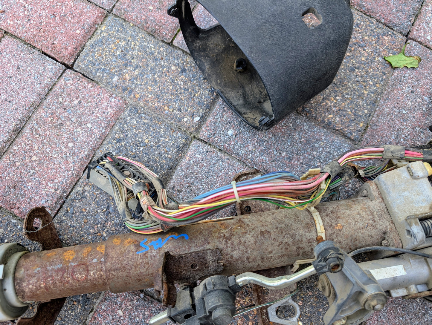A close-up of a General Motors OEM Steering Column w/Tilt, Cruise Control, and Column Shift for 1998-2005 Chevy S10 Blazer, showing colorful wires and connectors outdoors on a brick pavement with the black cover detached.