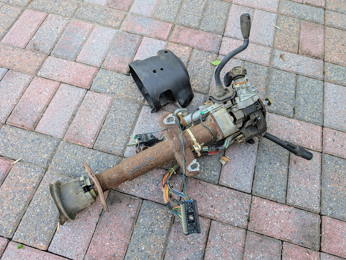 A General Motors OEM Steering Column with Tilt, Cruise Control, and Column Shift for 1998-2005 Chevy S10 Blazer lies on brick pavement next to its black plastic cover, showing exposed wiring and attached components.
