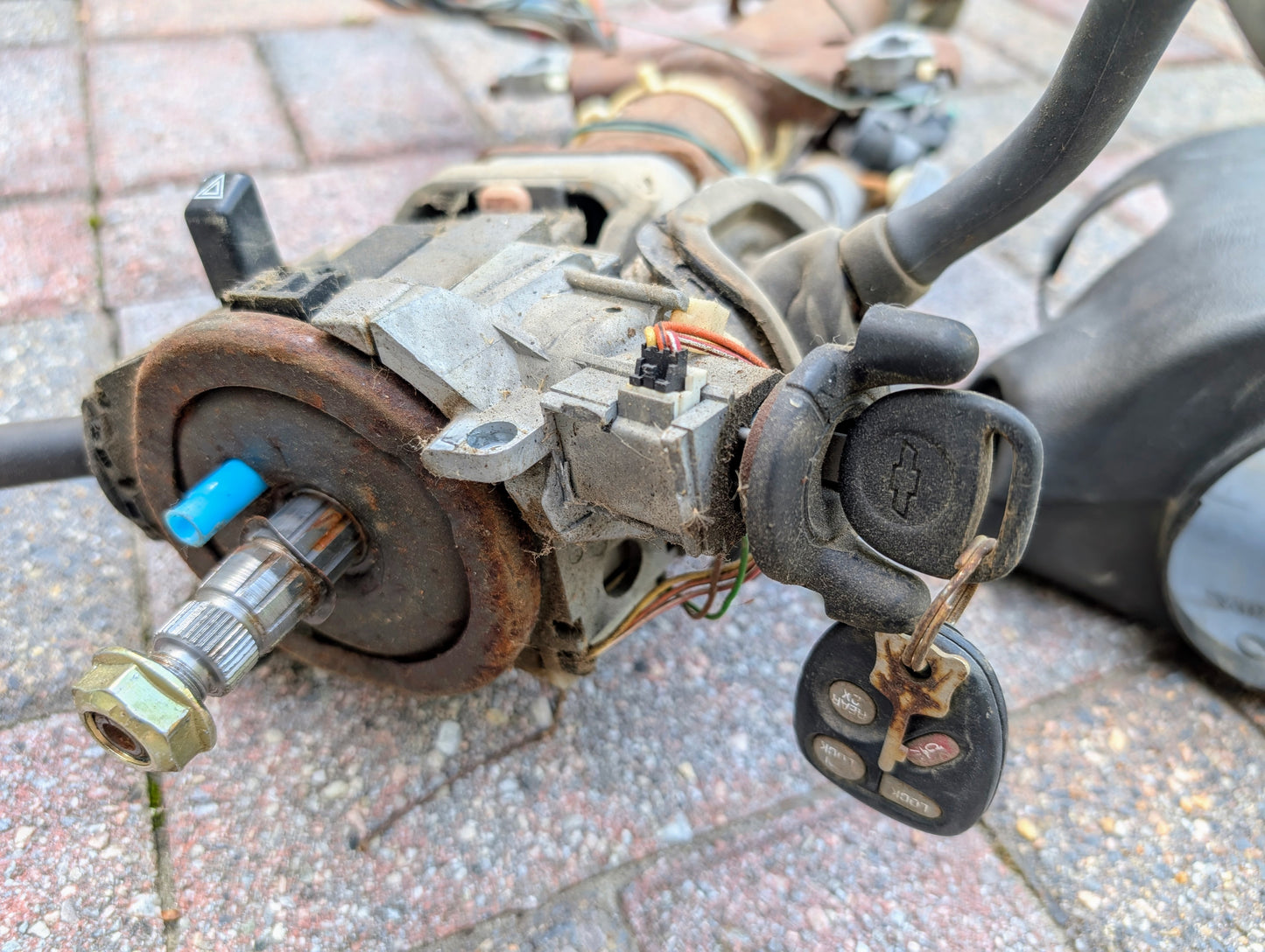 A close-up of a General Motors OEM Steering Column with tilt, cruise control, and column shift for 1998-2005 Chevy S10 Blazer shows the ignition with key and remote fob inserted; metal parts appear worn, rusted, and have exposed wires.