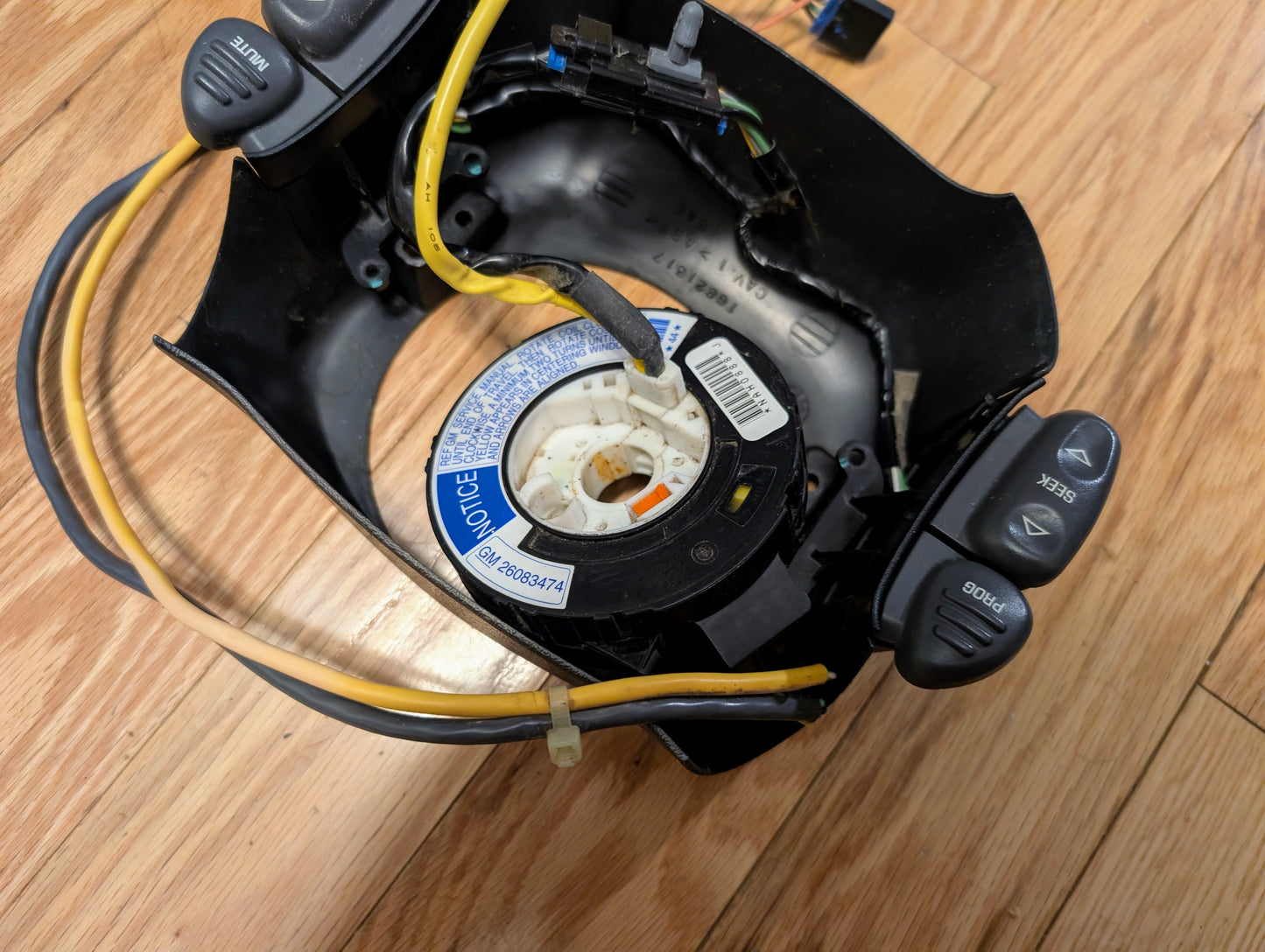 A General Motors KIT Steering Wheel Radio Controls & Clock Spring for 2000-2003 Chevy S10, Blazer, and more is shown disassembled on a wooden floor, with wiring, radio control buttons, inner mechanism, and caution label visible.