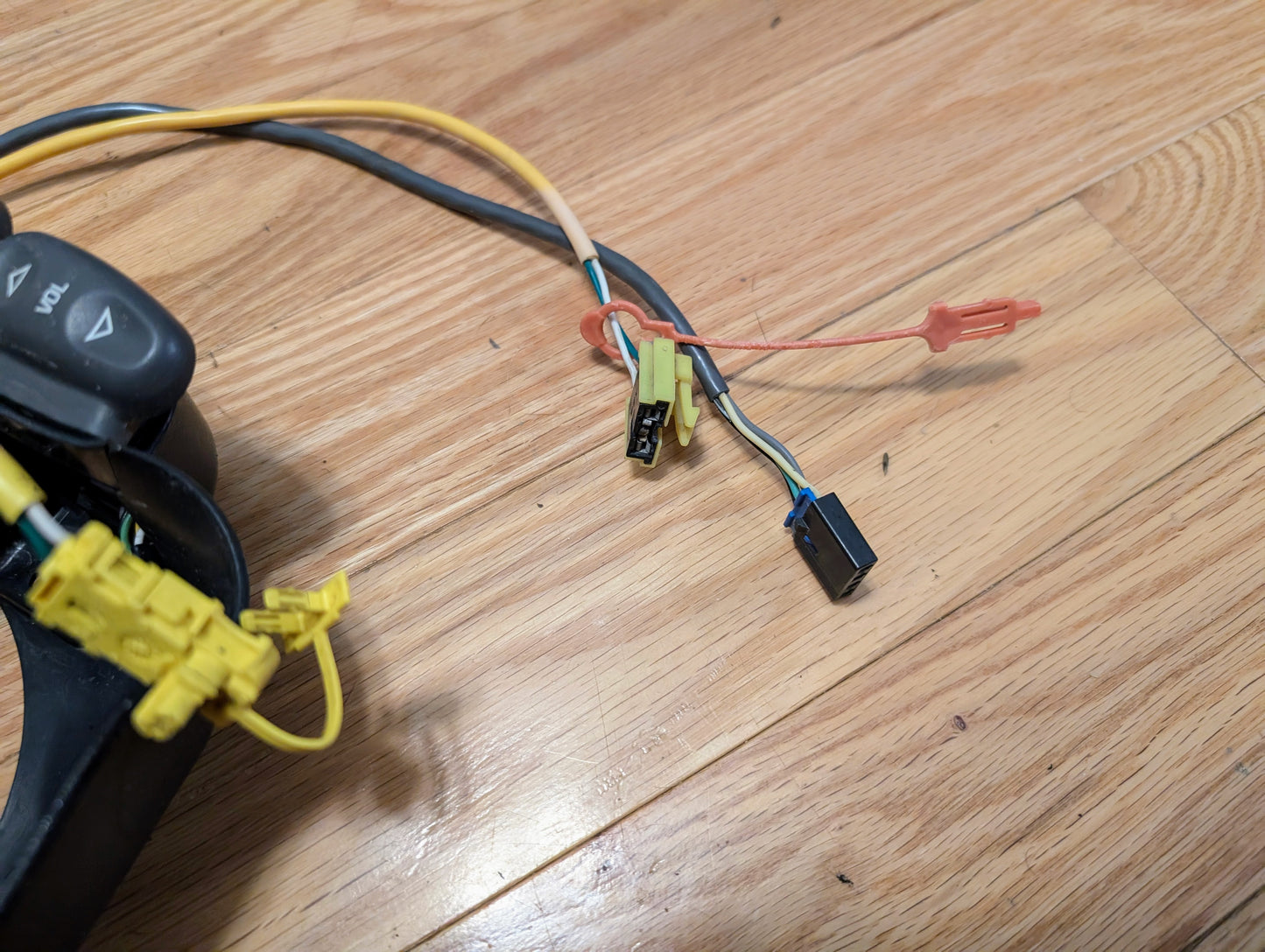 A close-up of disconnected wiring harnesses and connectors from the General Motors KIT Steering Wheel Radio Controls & Clock Spring for 2000-2003 Chevy S10, Blazer, and more, with an orange clip and part of the controls on a wooden floor.