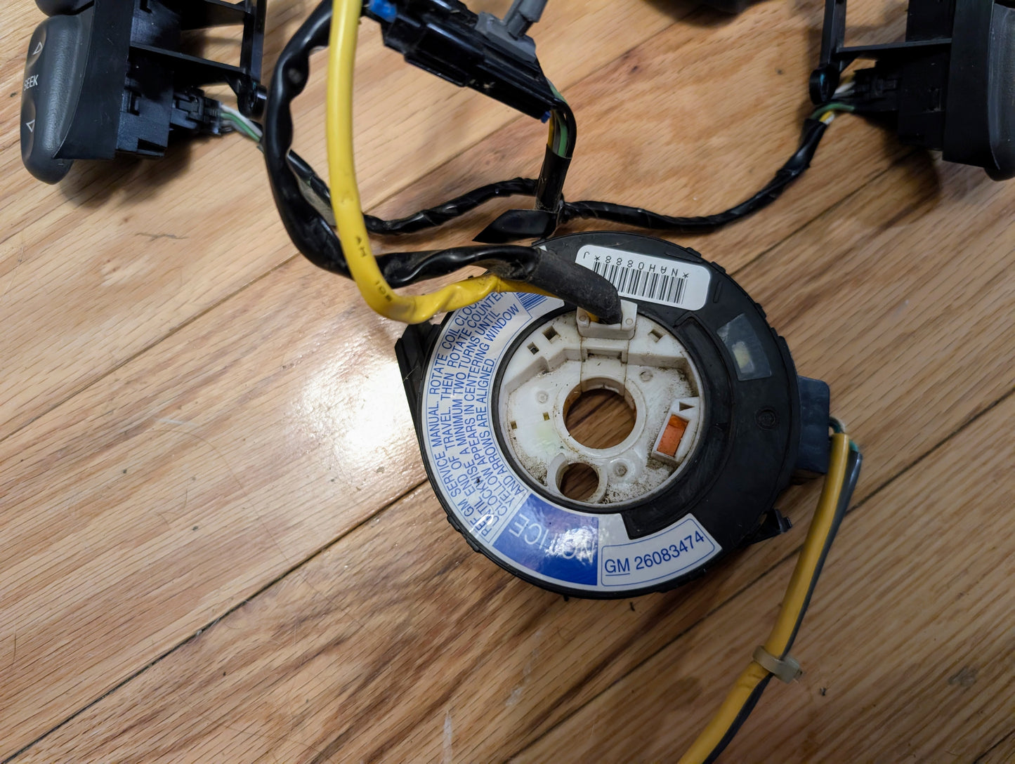 A close-up of a General Motors KIT Steering Wheel Radio Controls & Clock Spring for 2000-2003 Chevy S10, Blazer, and more, showing a circular part labeled GM 26093471 with wires attached and blue warning text on a wooden floor.
