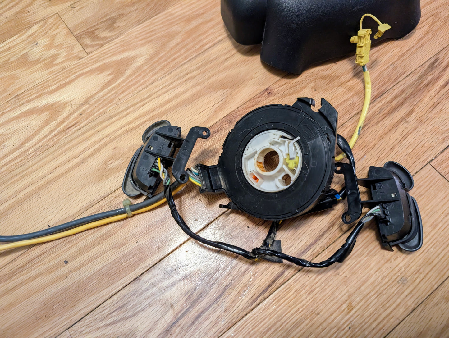 A General Motors KIT Steering Wheel Radio Controls & Clock Spring for 2000-2003 Chevy S10 and Blazer, with wiring and connectors, is shown on a wooden floor next to a separated plastic steering column cover.