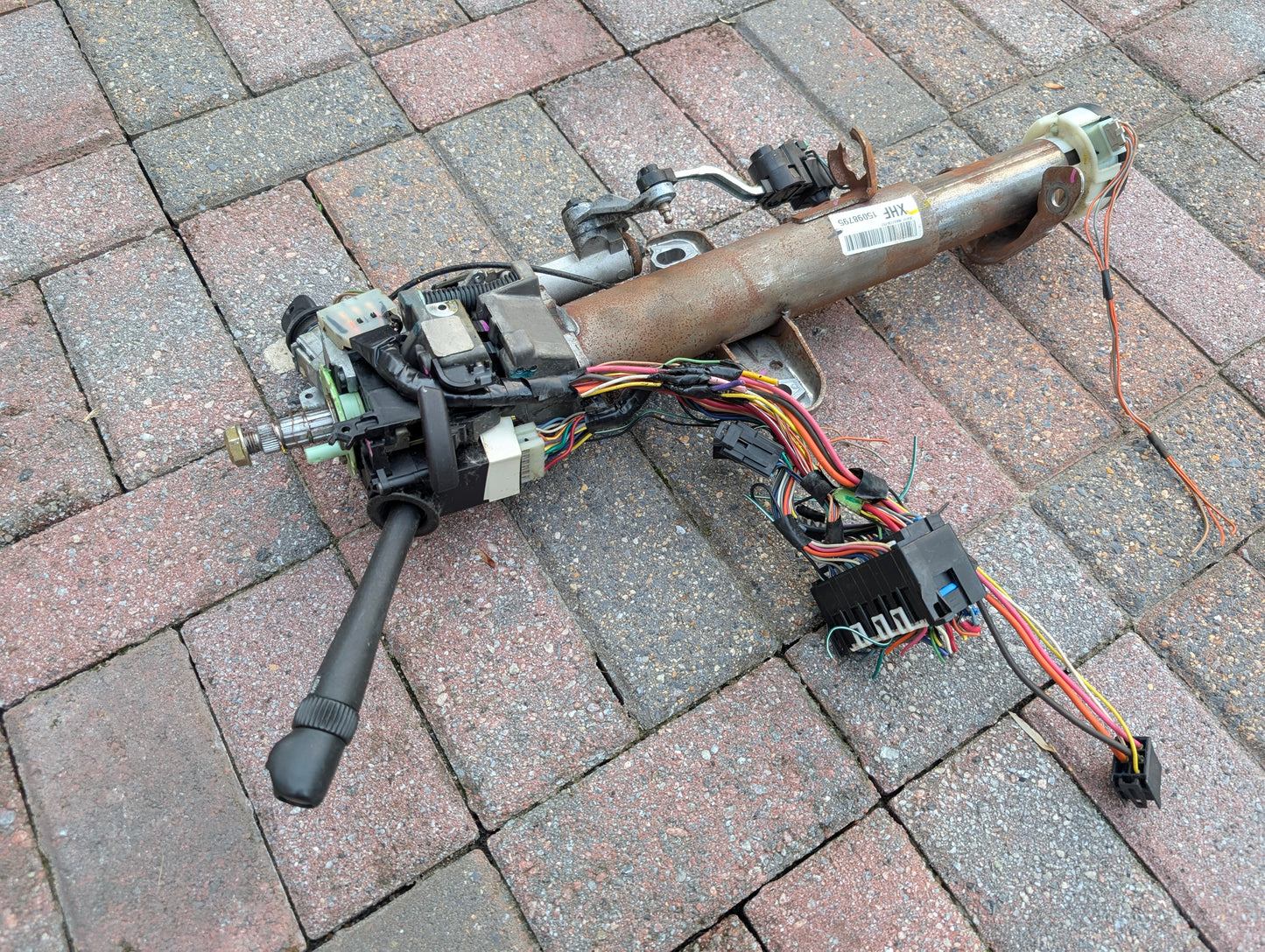 A General Motors OEM Steering Column w/KEY for 2000-2002 GM Full Size Trucks, featuring wiring and control stalks plus an automatic transmission shifter, is shown disassembled on a brick paved surface.