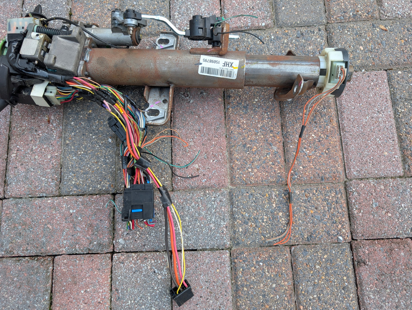 A General Motors OEM Steering Column w/KEY for 2000-2002 GM Full Size Trucks, featuring a wiring harness, connectors, and modules with visible colored wires, rests on a brick-paved ground.
