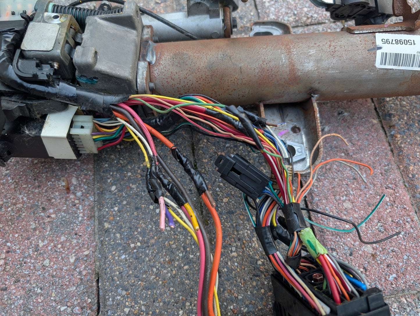 A close-up of colorful, tangled automotive wires connected to metal parts, likely from a General Motors OEM Steering Column w/KEY for 2000-2002 GM Full Size Trucks, some wires spliced with heat-shrink tubing, resting on textured pavement.