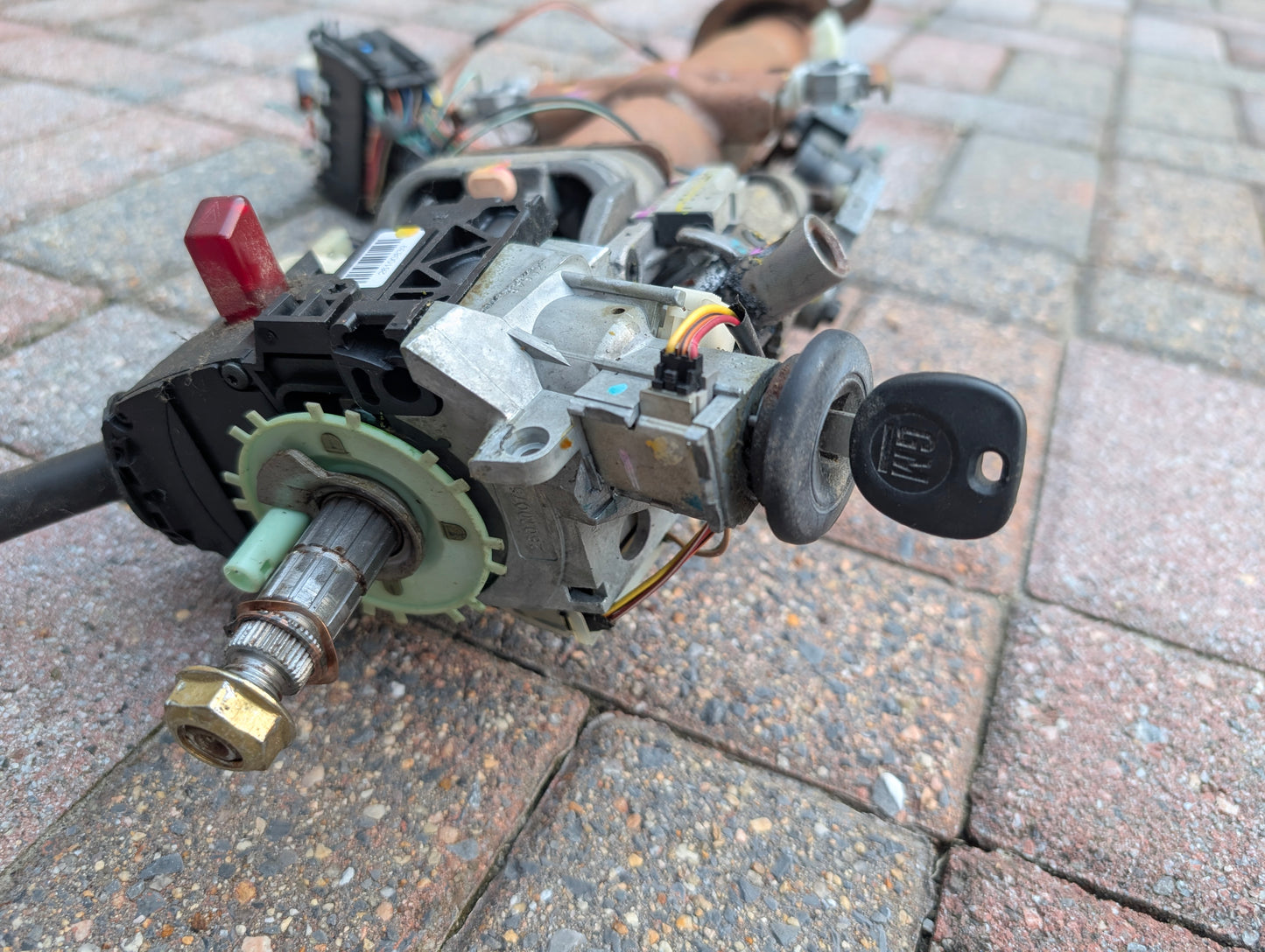 A close-up of a General Motors OEM Steering Column w/KEY for 2000-2002 GM Full Size Trucks shows the ignition switch assembly with inserted key, exposed wires, bolts, and connectors on a paved surface.