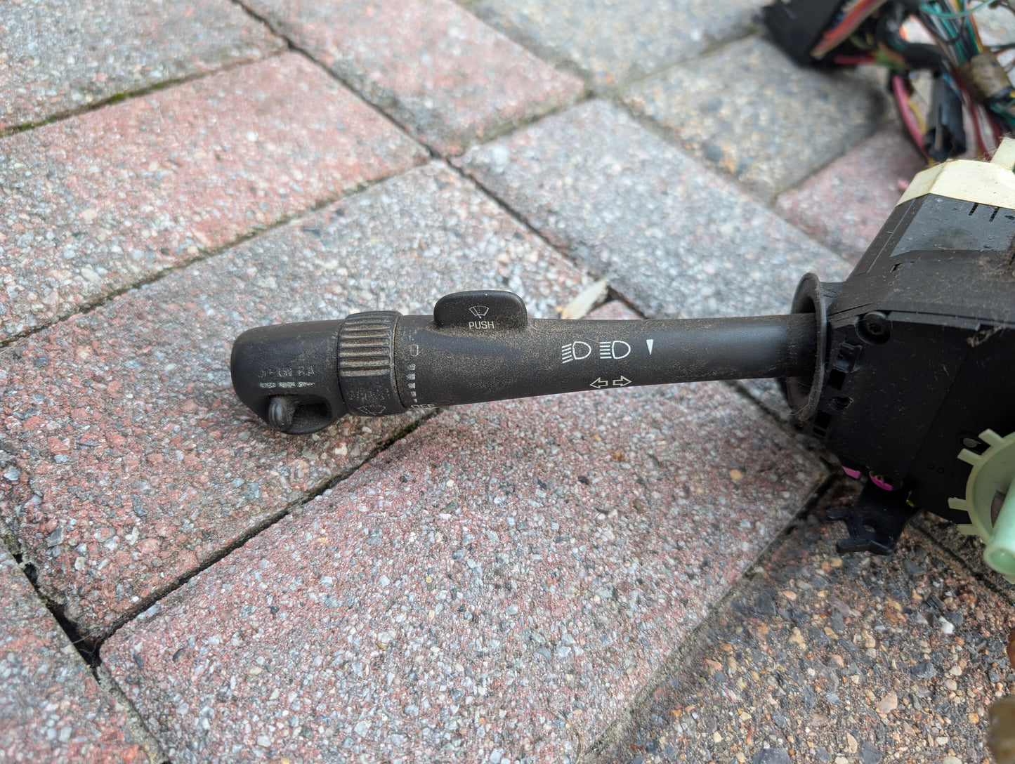 A close-up of a General Motors OEM Steering Column w/KEY for 2000-2002 GM Full Size Trucks rests on a textured red and gray brick surface, with some car wiring and connectors partially visible in the background.