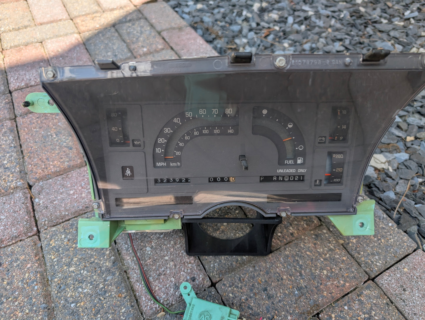 A General Motors OEM Instrument Cluster with Mechanical Speedo & PRNDL for 1986-1989 Chevy S10 Blazer is shown outdoors, featuring visible wiring and a clear casing displaying the speedometer, fuel gauge, odometer, and warning lights.