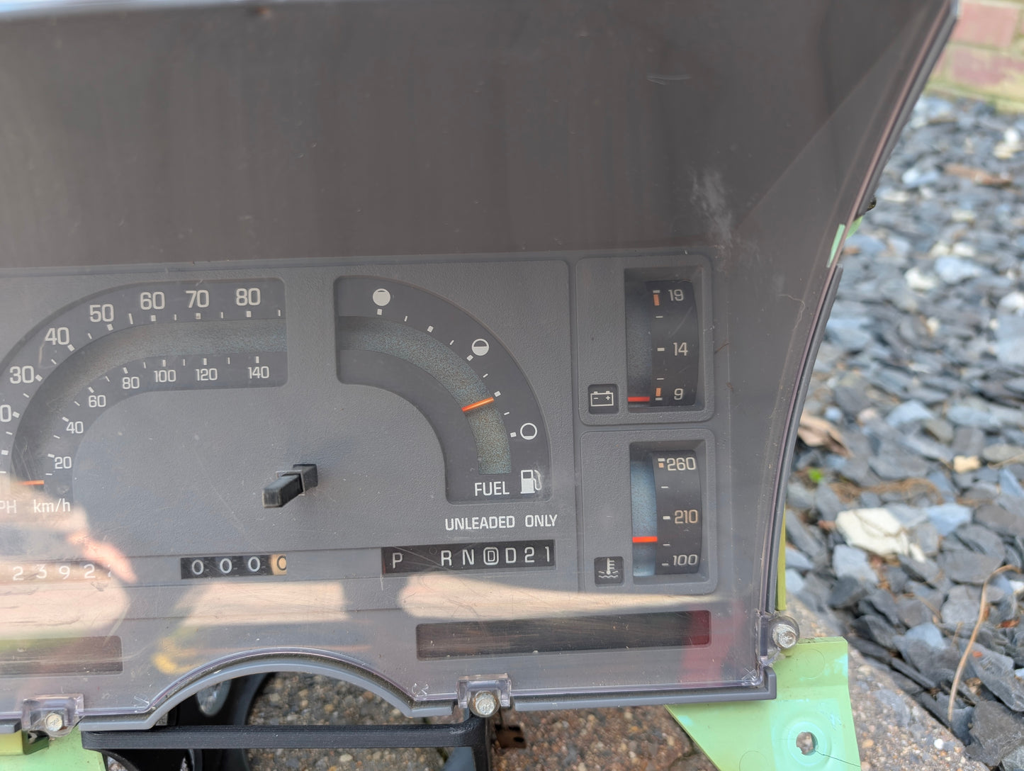 Close-up of the General Motors OEM Instrument Cluster with Mechanical Speedo & PRNDL for 1986-1989 Chevy S10 Blazer, showing speedometer, fuel and temperature gauges, gear indicator, and warning lights. Background includes gravel and a green vehicle panel.