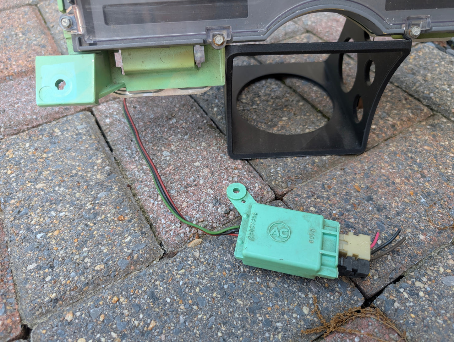 A green electrical connector with wires is attached to a General Motors OEM Instrument Cluster with Mechanical Speedo & PRNDL for 1986-1989 Chevy S10 Blazer, which is resting on a gray and brown brick surface.