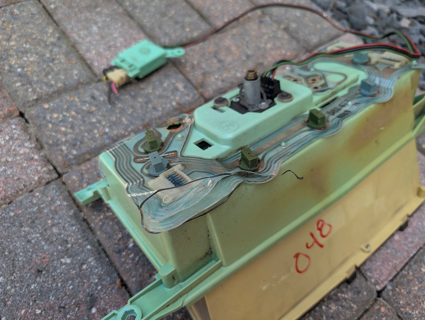A close-up of a General Motors OEM Instrument Cluster with Mechanical Speedo & PRNDL for 1986-1989 Chevy S10 Blazer shows wires attached, the number 048 in red on the yellow base, and another component in the background on a brick surface.