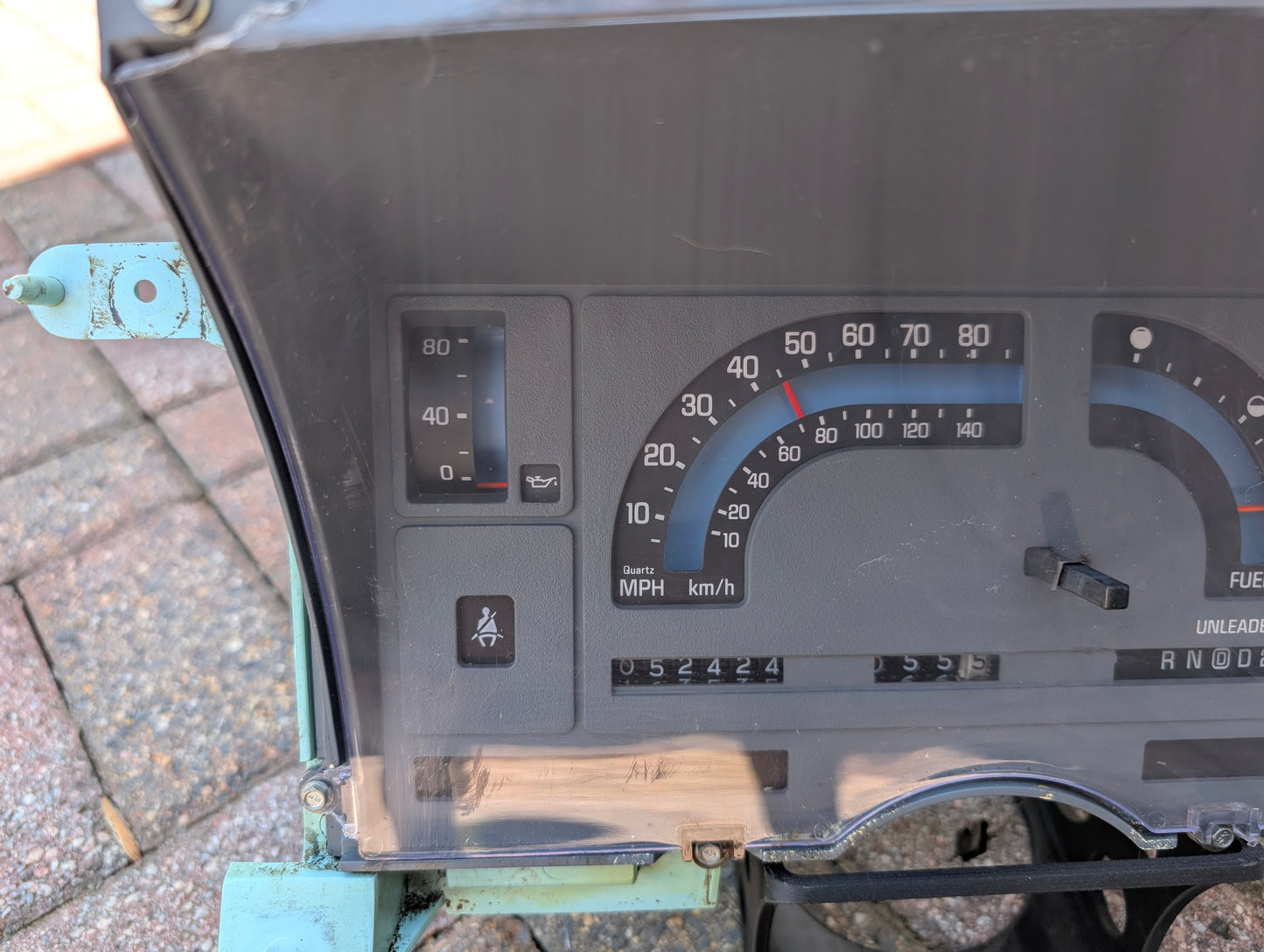 Close-up of a General Motors Low Mile 52K OEM Instrument Cluster with PRNDL for 1986-1994 Chevy S10 Blazer, showing speedometer, odometer reading 052424, fuel and oil gauges. Partially transparent dash reveals bricks in background.