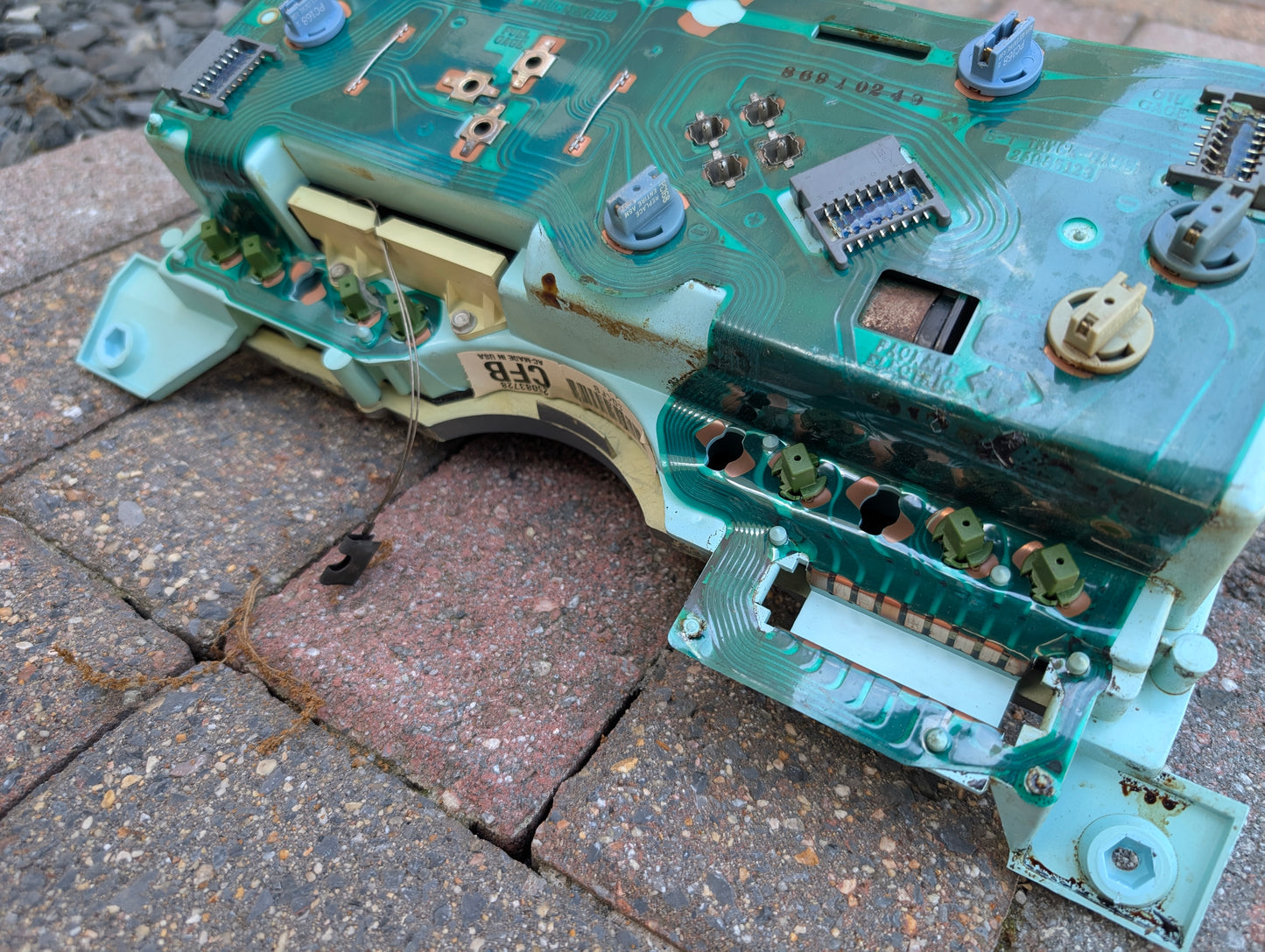 A close-up of a General Motors Low Mile 52K OEM Instrument Cluster with PRNDL for 1986-1994 Chevy S10 Blazer, showing weathered electronic components and signs of rust or corrosion, lying on outdoor bricks.