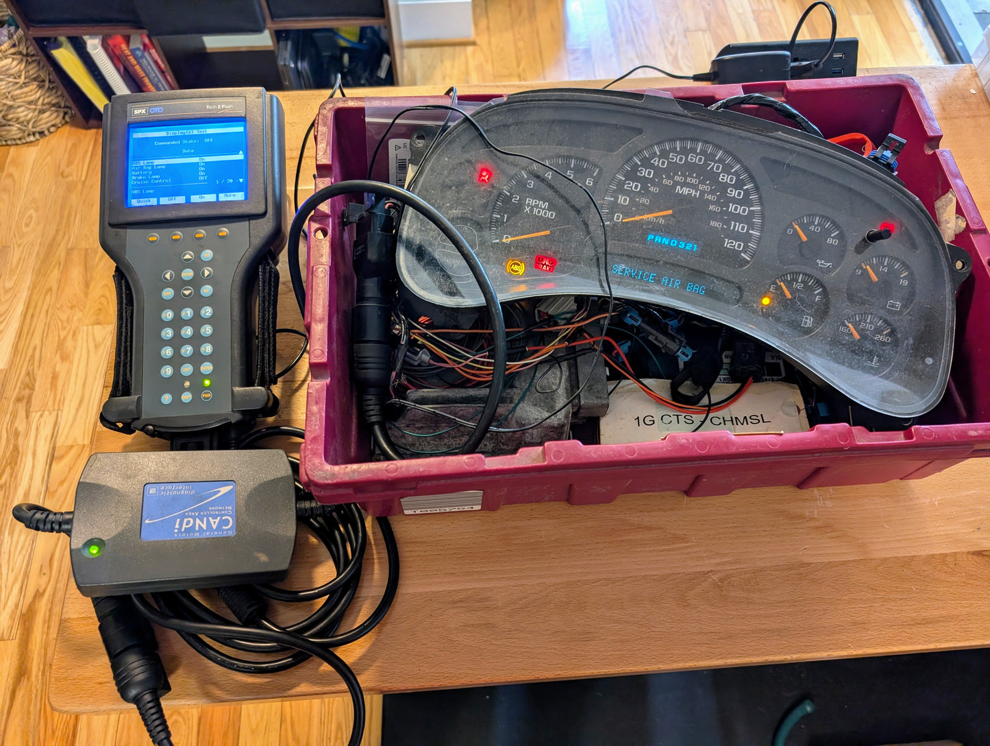 A Vetronix TESTED Tech 2 Automotive Diagnostic Tool & Accessories for 1991-2013 GM Vehicles is wired to a disassembled GM dashboard cluster in a plastic bin on a wooden table, with wiring and electronics visible.