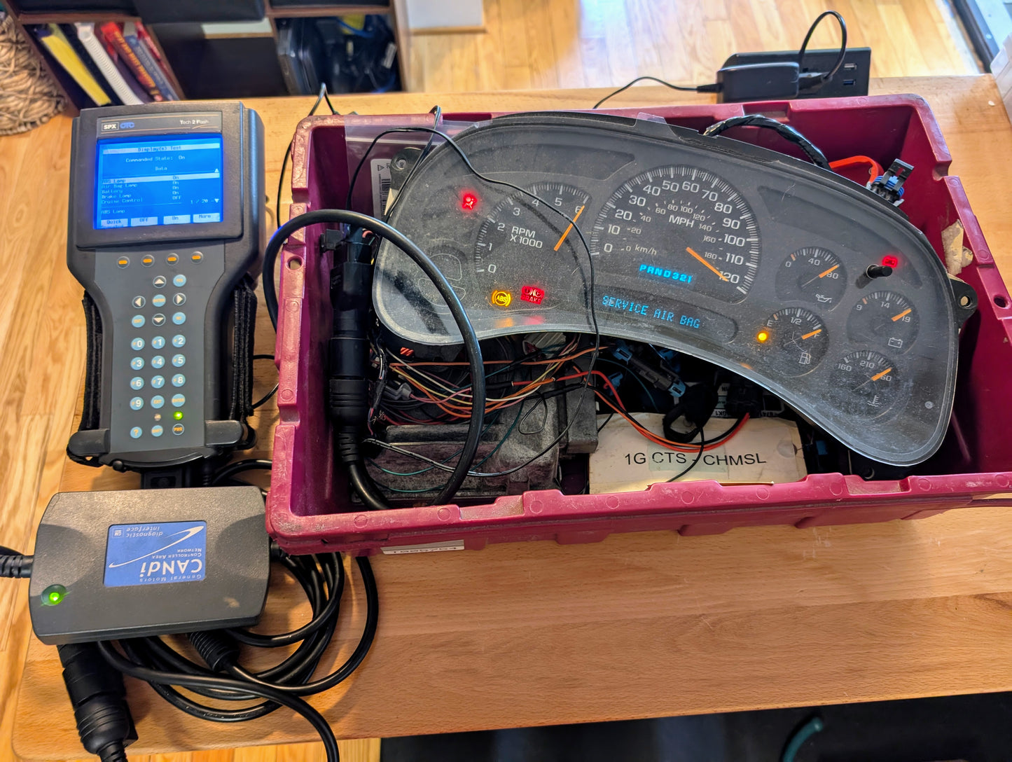 The Vetronix TESTED Tech 2 Automotive Diagnostic Tool & Accessories for 1991-2013 GM Vehicles connects via cables to a dashboard instrument cluster, showing illuminated indicator lights—perfect for diagnosing GM cars.