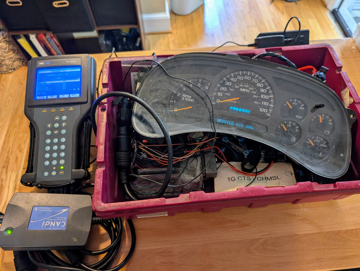 A Vetronix TESTED Tech 2 Automotive Diagnostic Tool & Accessories for 1991-2013 GM Vehicles is connected by cables to a car dashboard instrument cluster in a red plastic box on a wooden table, used for testing or repair.