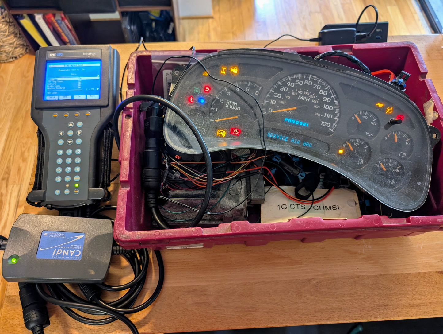 A Vetronix TESTED Tech 2 Automotive Diagnostic Tool & Accessories for 1991-2013 GM Vehicles is connected to a dashboard cluster, both set up with visible wiring in a plastic bin on a wooden table indoors.