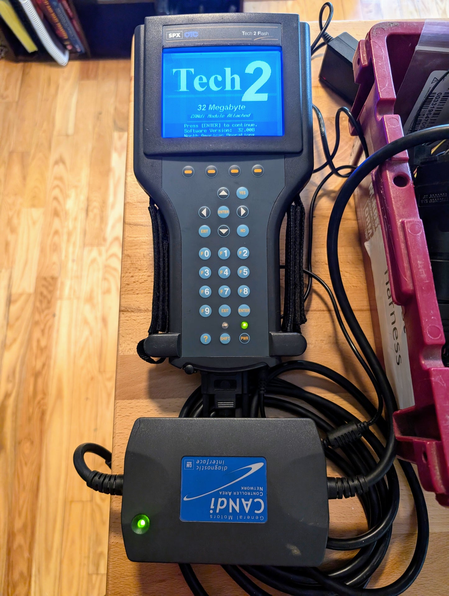 A Vetronix TESTED Tech 2 Automotive Diagnostic Tool & Accessories for 1991-2013 GM vehicles, featuring a blue screen and CANDi module, are shown on a wooden floor beside a red storage bin—perfect for GM diagnostics.