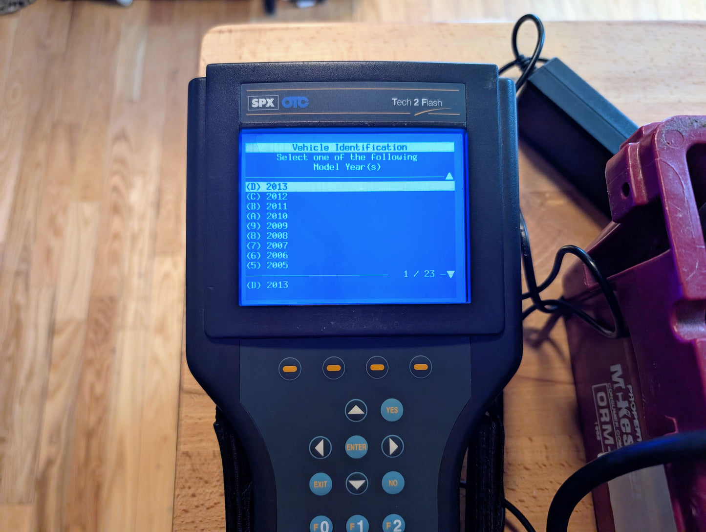 The Vetronix TESTED Tech 2 Automotive Diagnostic Tool & Accessories for 1991-2013 GM vehicles shows a blue menu for year selection. The handheld OBD2 scanner, with cables attached, sits on a wooden surface.