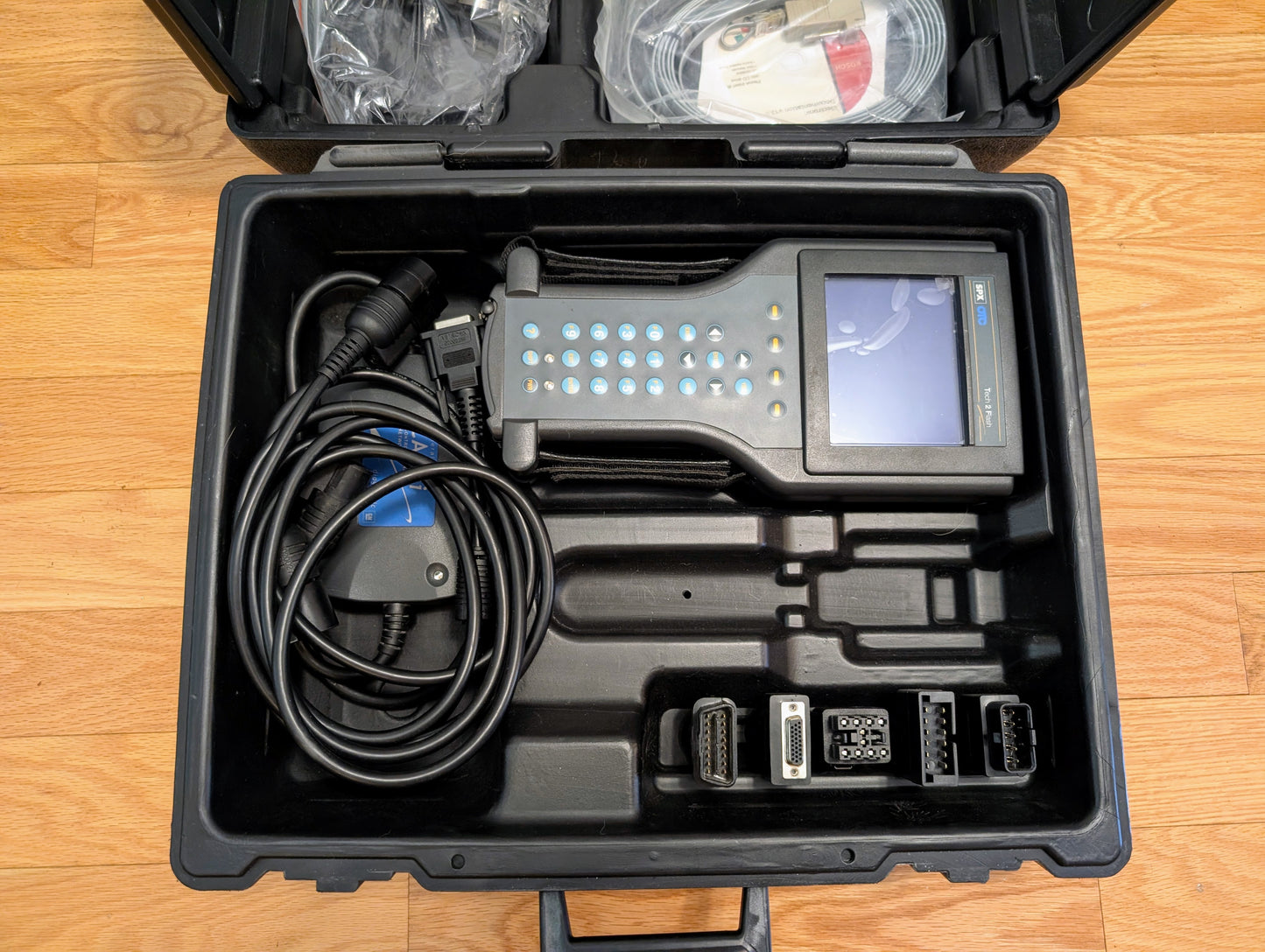A black Vetronix case holds the TESTED Tech 2 Automotive Diagnostic Tool & Accessories for 1991-2013 GM vehicles, with keypad, screen, cables, and adapters neatly organized. The OBD2 scanner is open on a wooden floor.