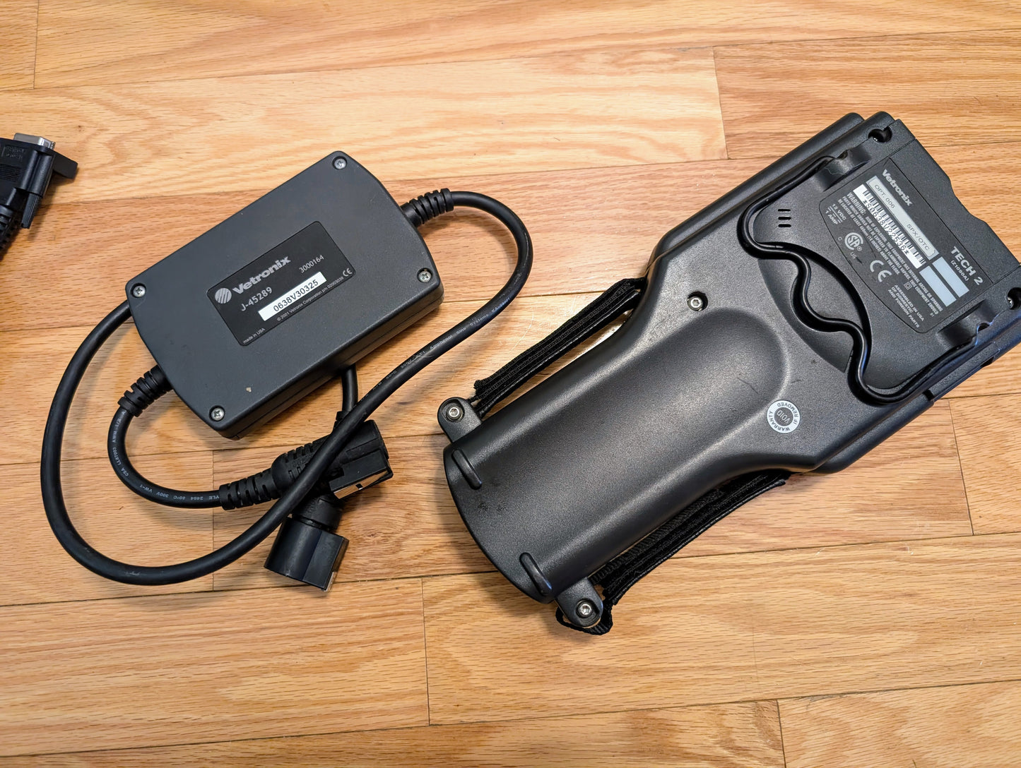 A Vetronix TESTED Tech 2 Automotive Diagnostic Tool & Accessories for 1991-2013 GM Vehicles, with a black lithium-ion battery charger and large labeled battery pack, is placed on a wooden floor. The charger is connected by a cable, showing clear brand and model labels.