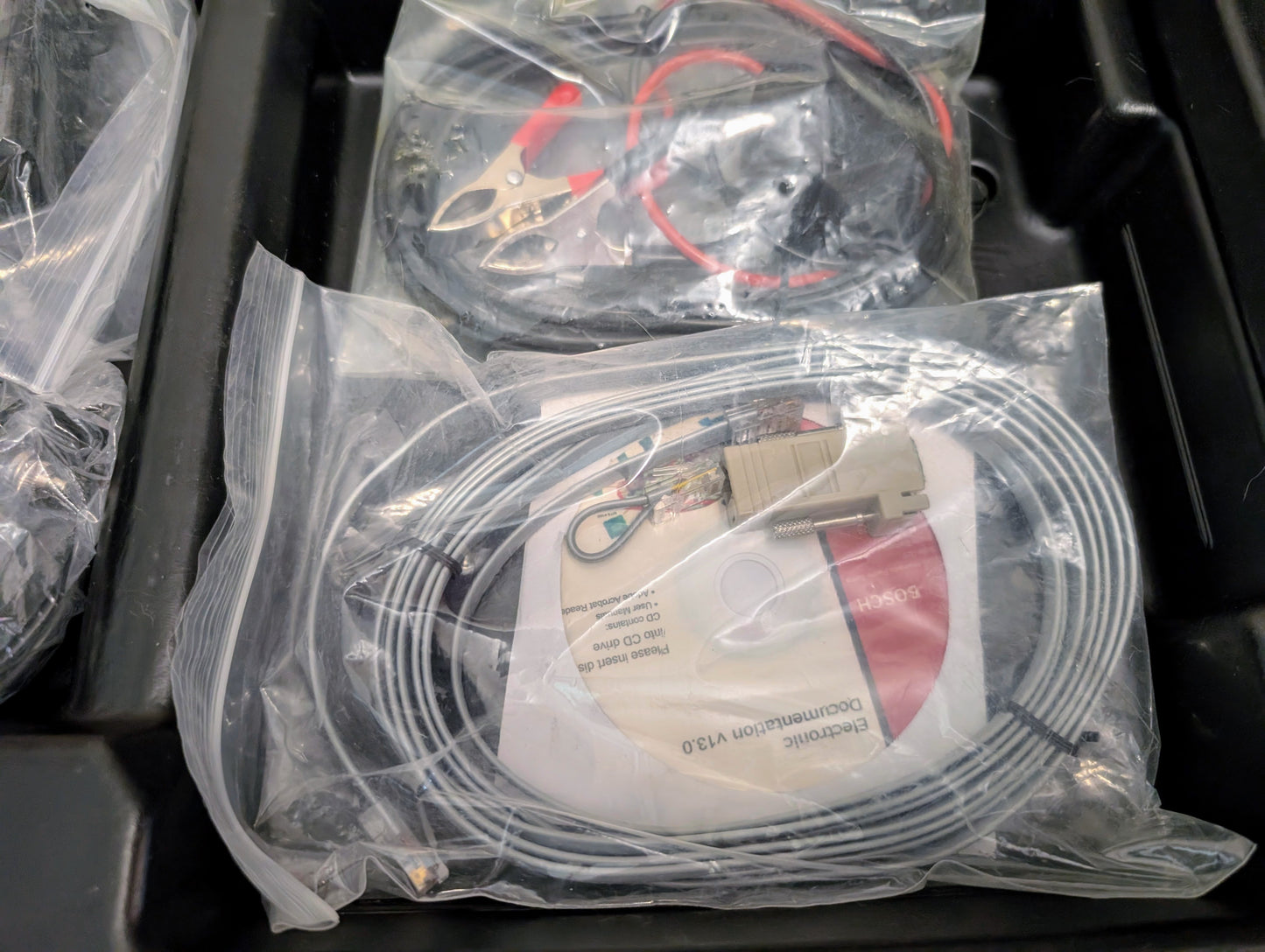A clear plastic bag in a black tray holds a coiled gray cable with connectors and a red and white instruction sheet. Another bag with alligator clips for the Vetronix TESTED Tech 2 Automotive Diagnostic Tool (1991-2013 GM) is partially visible above.
