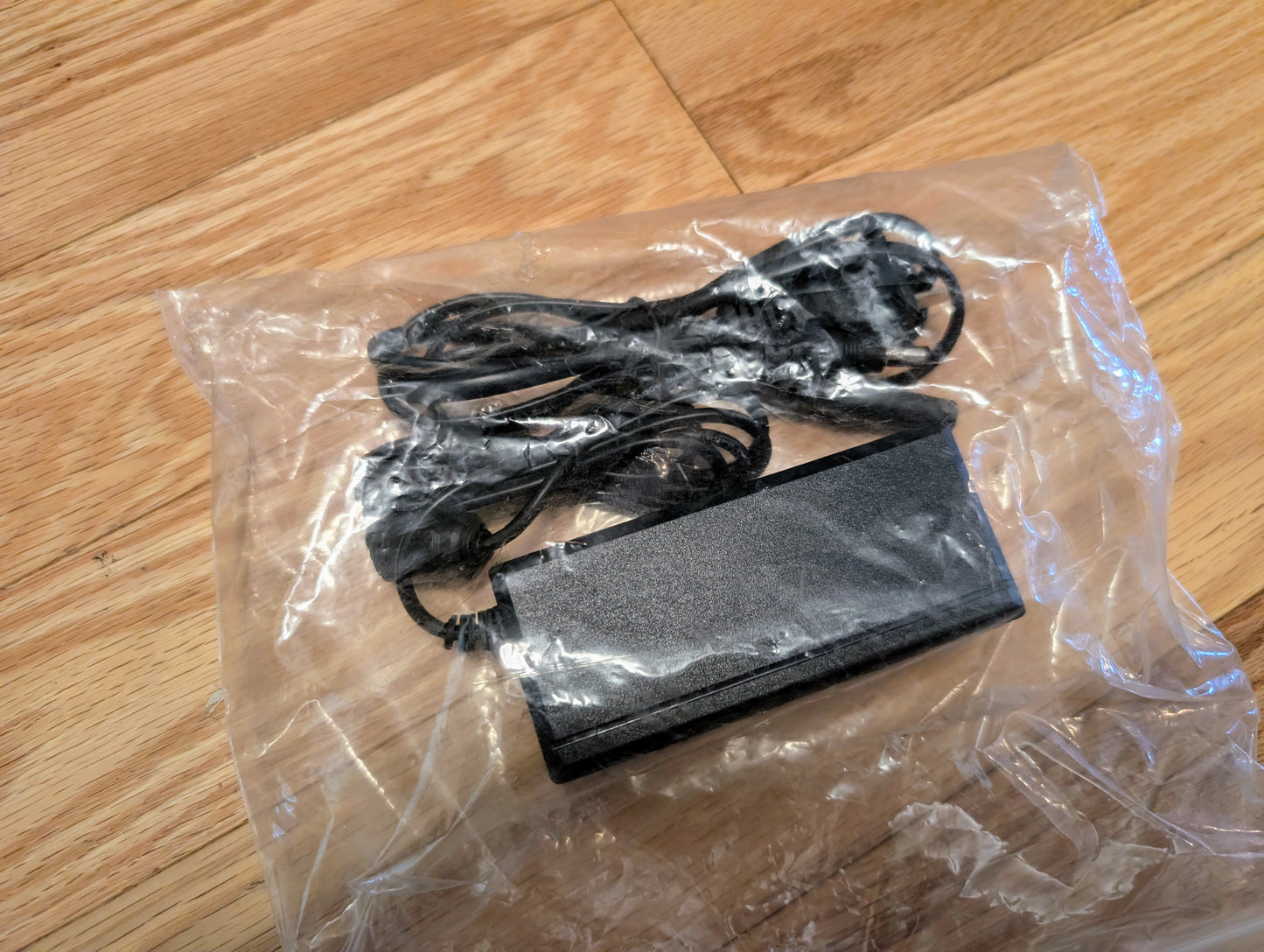 A black power adapter with a coiled cable, packaged in clear plastic, sits on a wooden floor—ideal for powering your Vetronix TESTED Tech 2 Automotive Diagnostic Tool & Accessories for 1991-2013 GM vehicles.