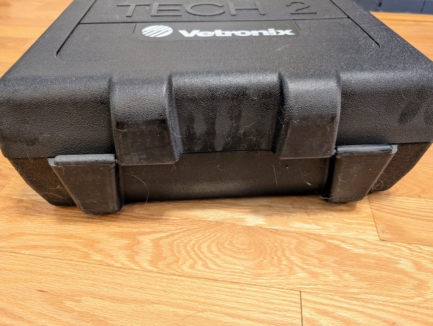 A black plastic carrying case labeled TESTED Tech 2 Automotive Diagnostic Tool & Accessories for 1991-2013 GM Vehicles by Vetronix sits on a wooden floor. The case has visible clasps and some dust or hair on its surface.