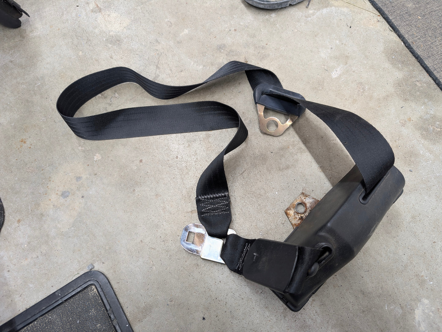 A General Motors NOS Passenger Seat Belt Retractor in Charcoal for 1982-1993 Chevy S10, Blazer, and more, with metal buckle and brackets, is partially extended and twisted near its casing on a concrete floor.