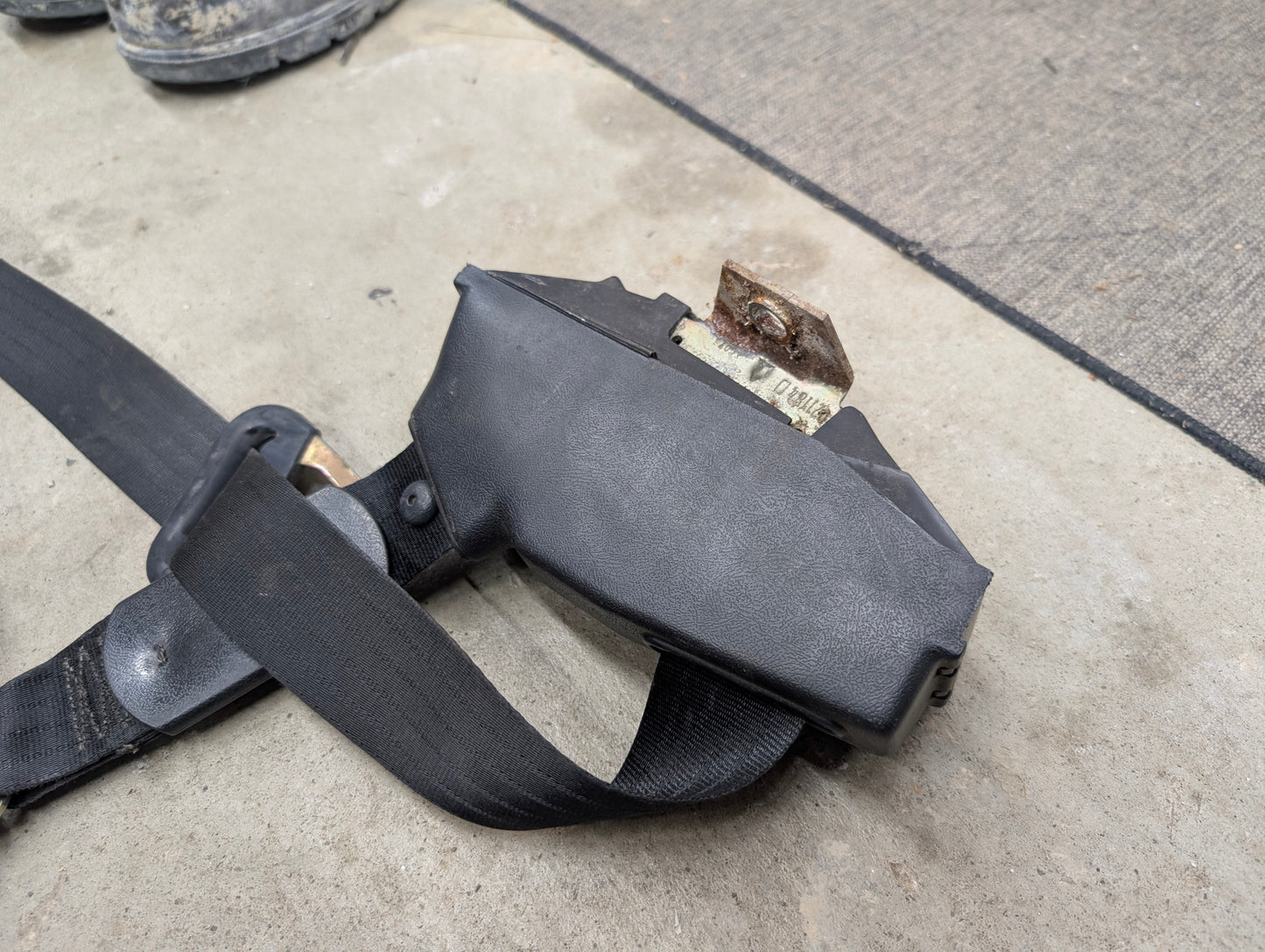 A NOS General Motors passenger seat belt retractor in charcoal for 1982-1993 Chevy S10, Blazer, and more, shown on a concrete surface.