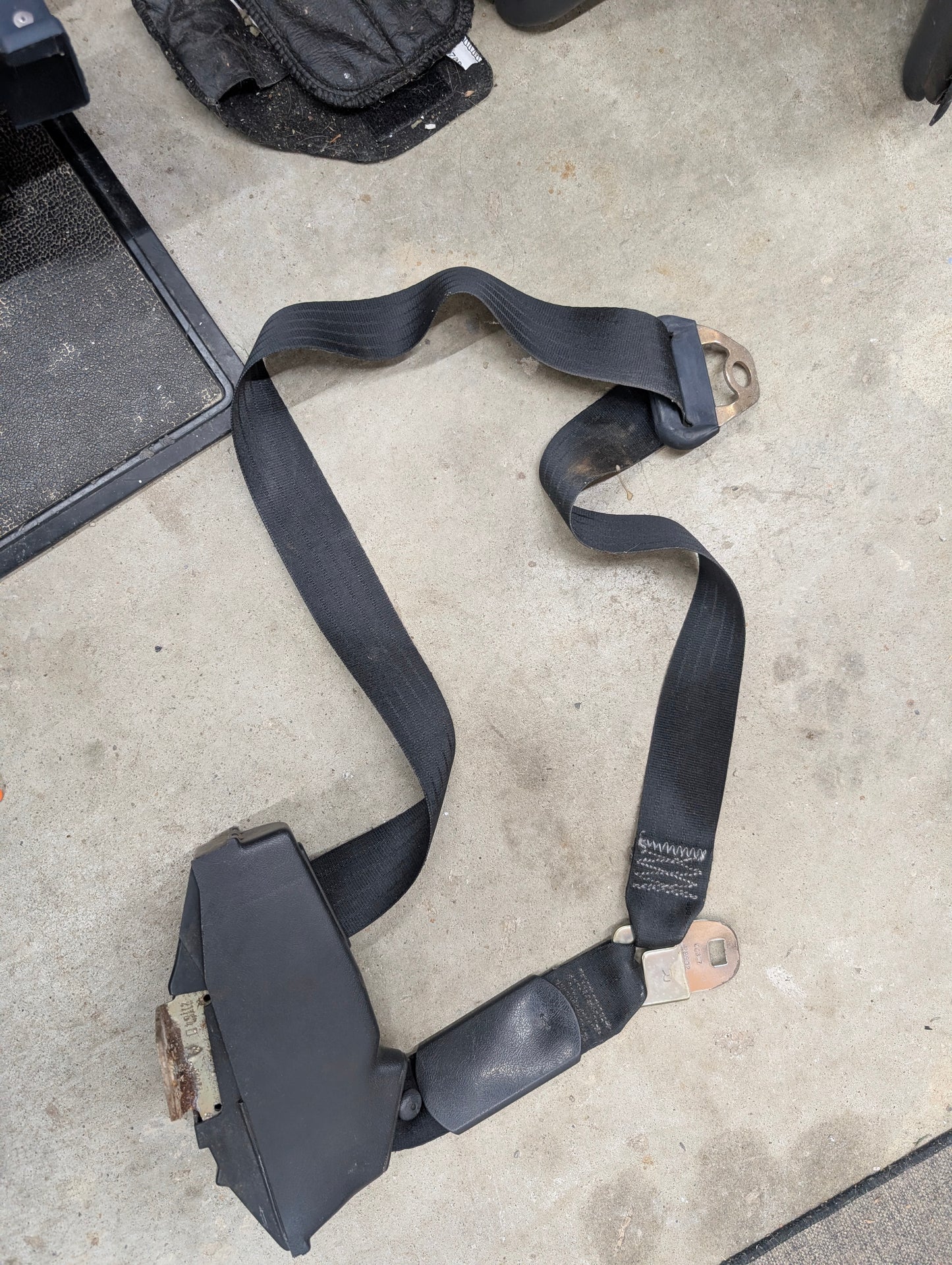 A NOS General Motors passenger seat belt retractor in charcoal, designed for 1982–1993 Chevy S10, Blazer, and more, lies on a concrete floor among various dark objects and equipment parts.