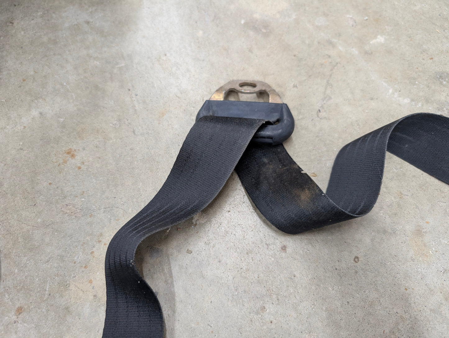 A General Motors NOS passenger seat belt retractor in charcoal for 1982–1993 Chevy S10, Blazer, and more rests on a gray concrete floor. It features a metal mounting bracket and fits vehicles with a charcoal interior.